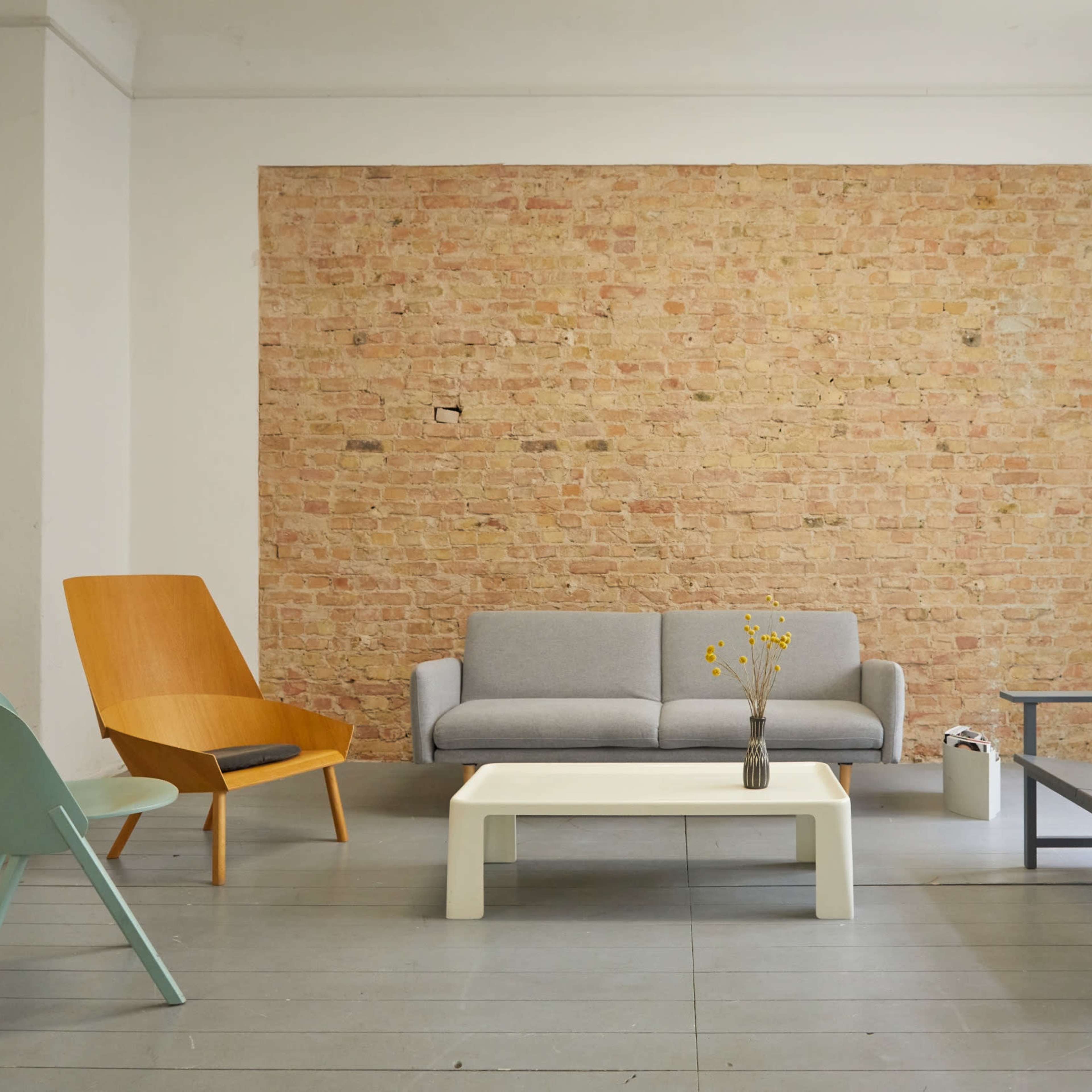 A minimalist living room with a gray sofa, an orange accent chair, a white coffee table, and a gray bench against a brick wall.
