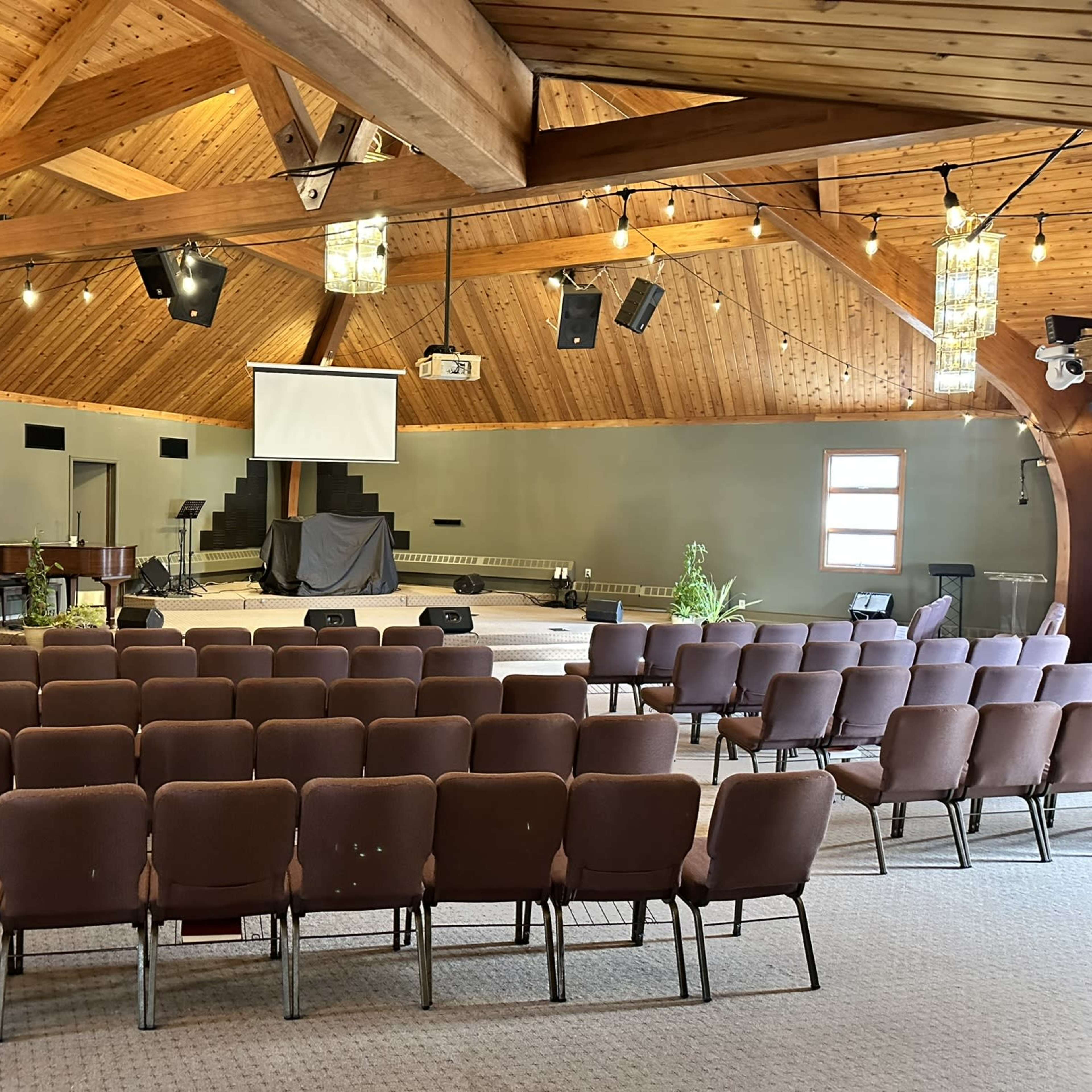 The interior of a church with wooden beams, rows of brown chairs facing a stage with a projector screen and piano.