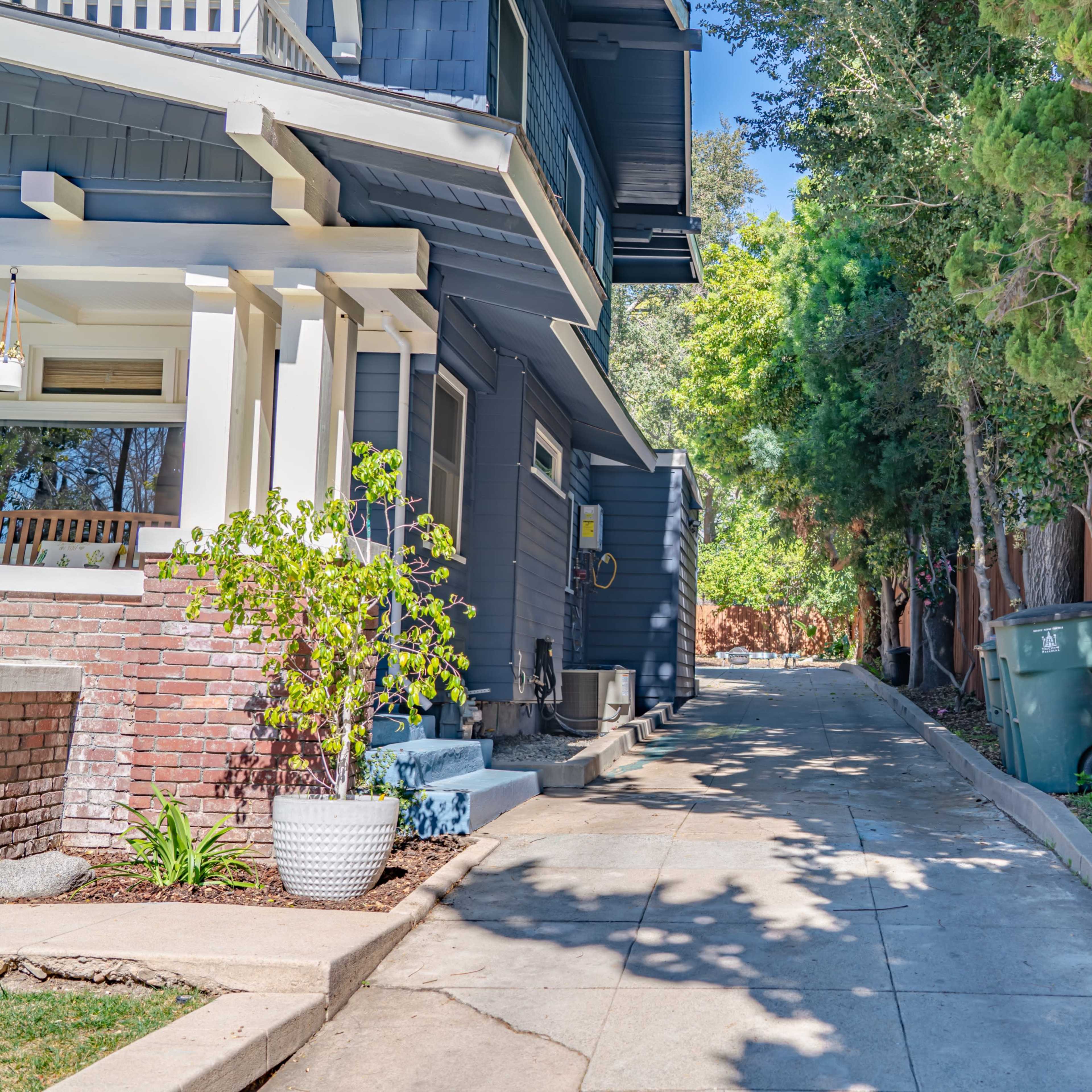 A narrow concrete pathway runs between a house and a fence, lined with trees and a green trash bin.