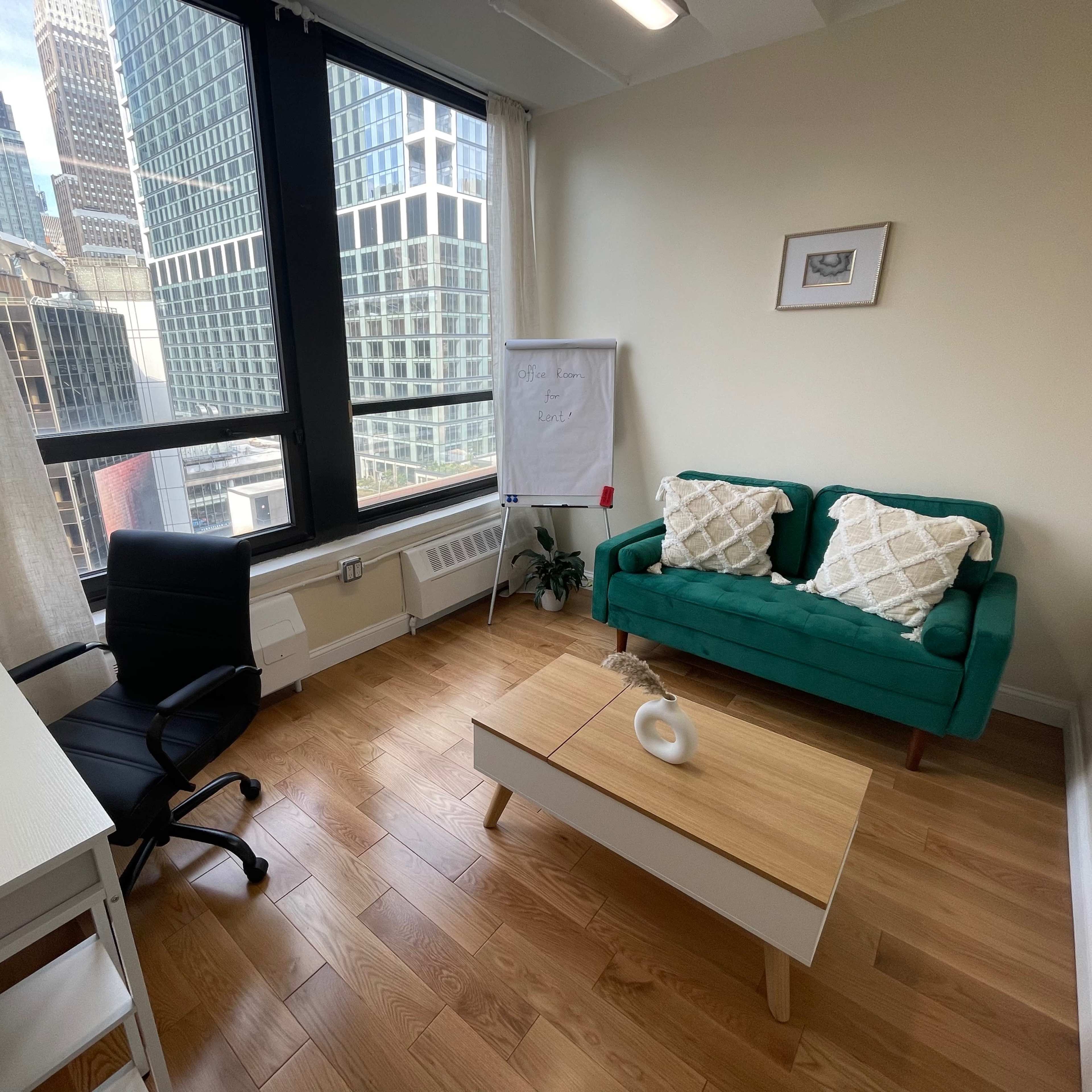 An office space features a green couch with patterned pillows, a wooden coffee table, a black chair, a white desk, and large windows overlooking a cityscape.