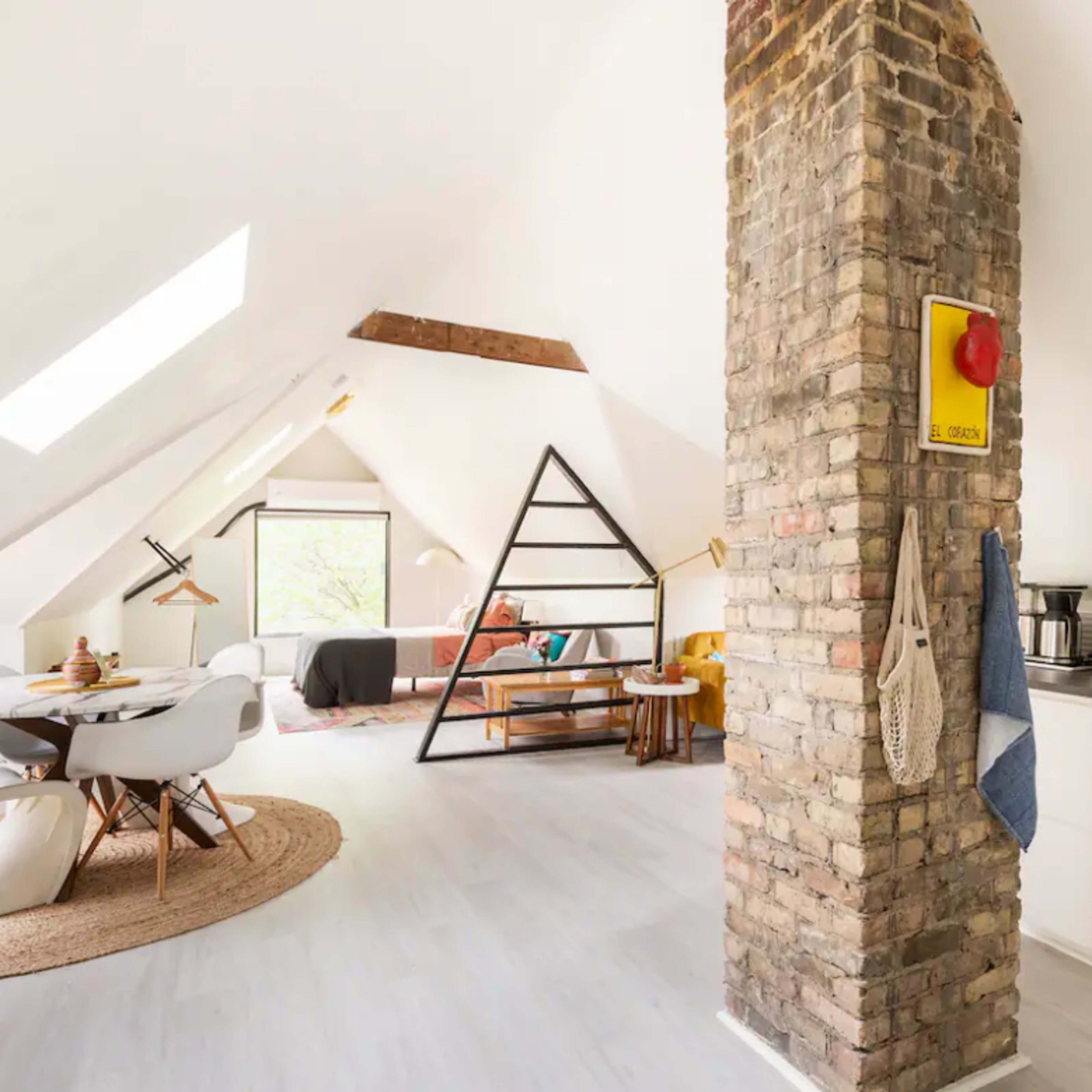 A modern attic apartment featuring a kitchen area, dining space with white chairs, and a cozy living area with a distinct brick column and skylights.