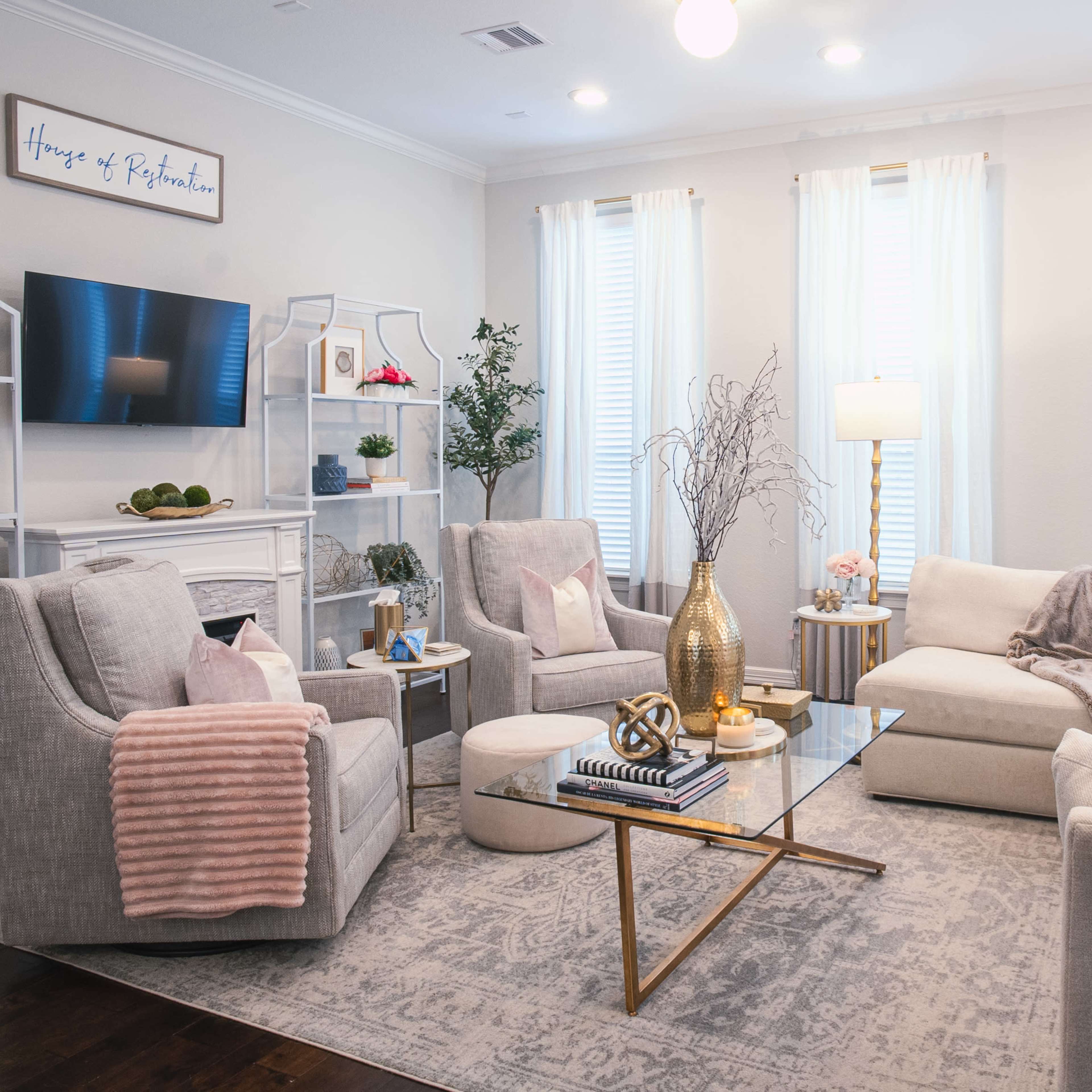 A modern living room features a cream-colored sectional sofa, two light gray armchairs, a glass coffee table, and a white bookshelf adorned with decorative items.