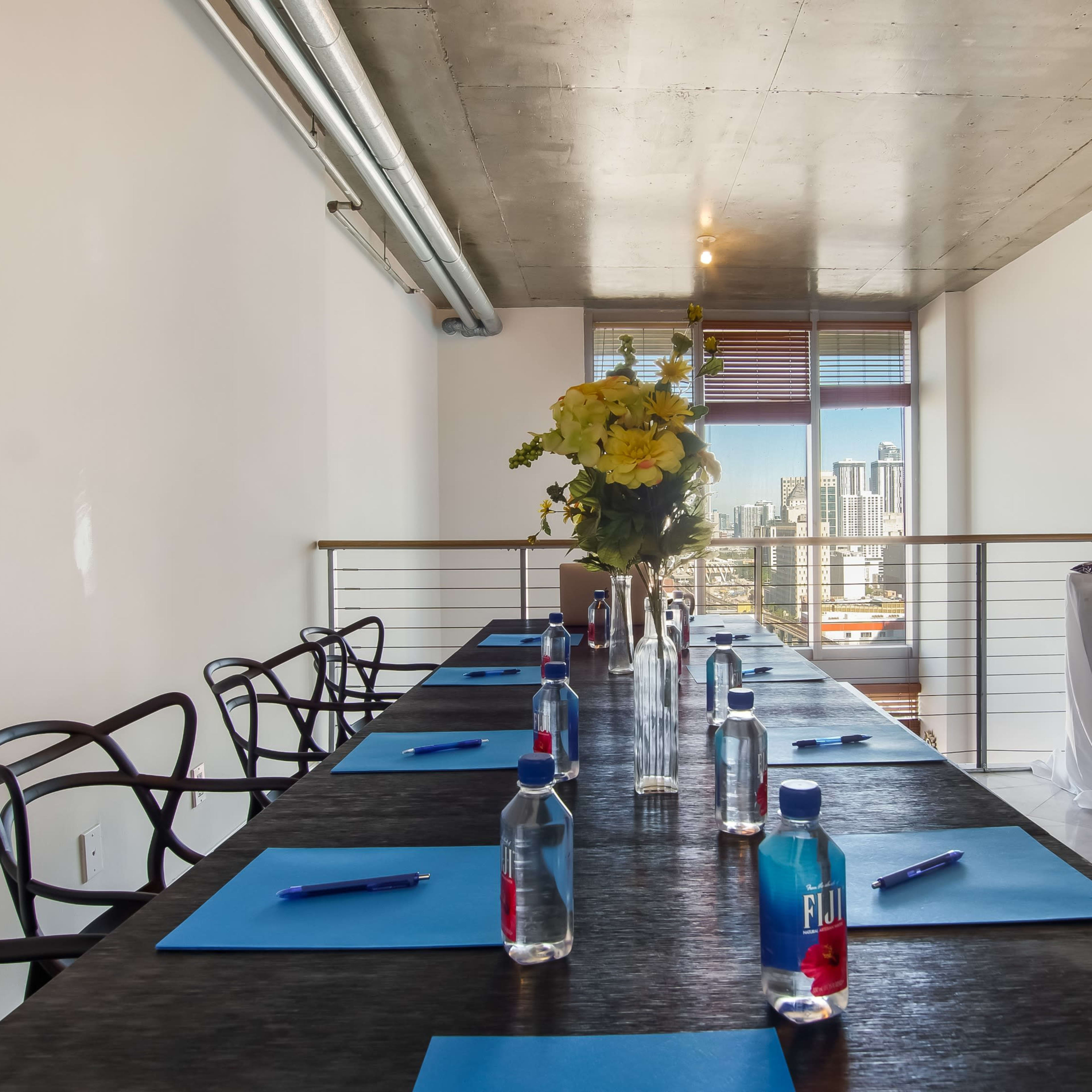 A long conference table is set with blue notepads and bottles of water, featuring a floral centerpiece, against a backdrop of city skyline views from a modern office space.