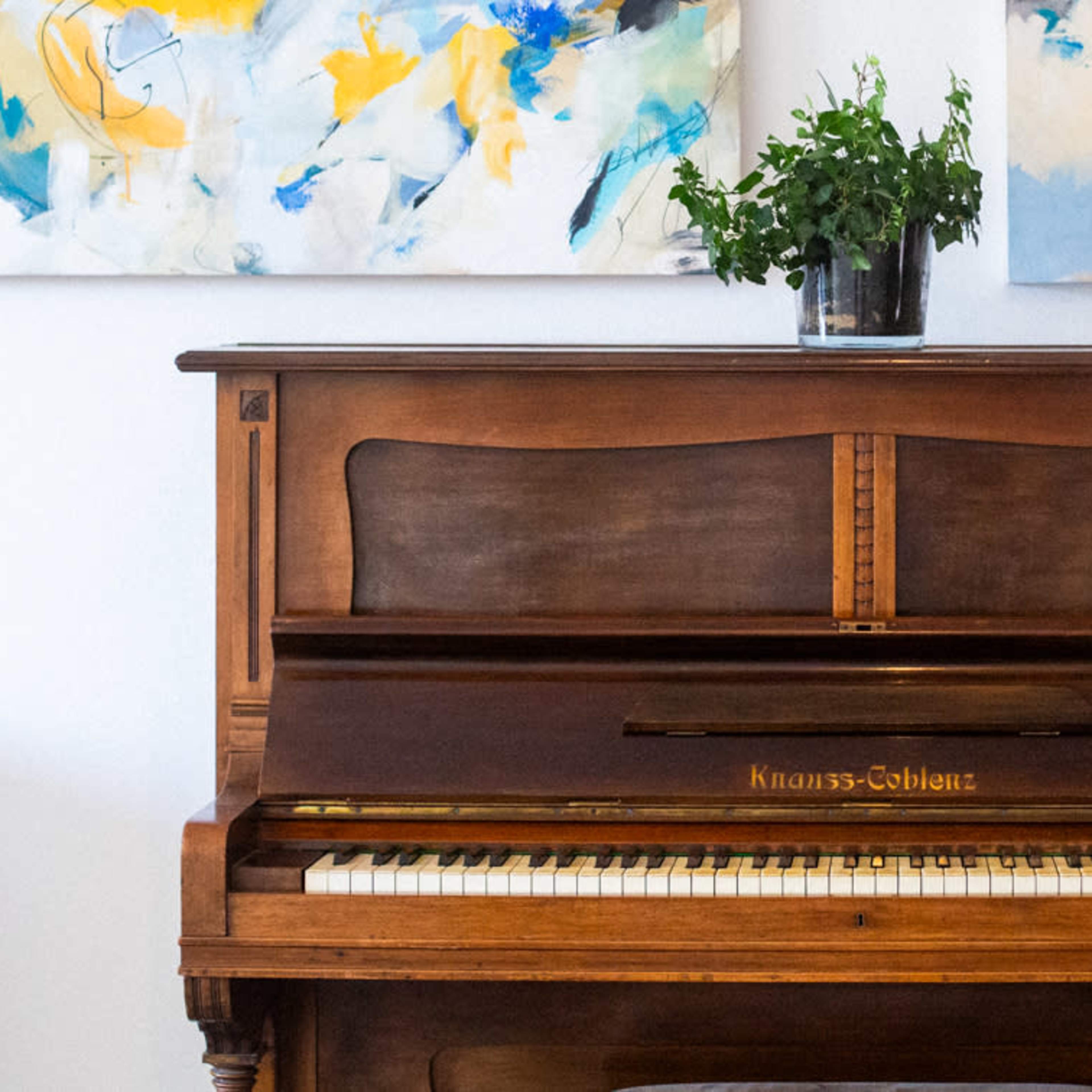 A vintage wooden piano is positioned between two abstract paintings, with a potted plant placed on top.