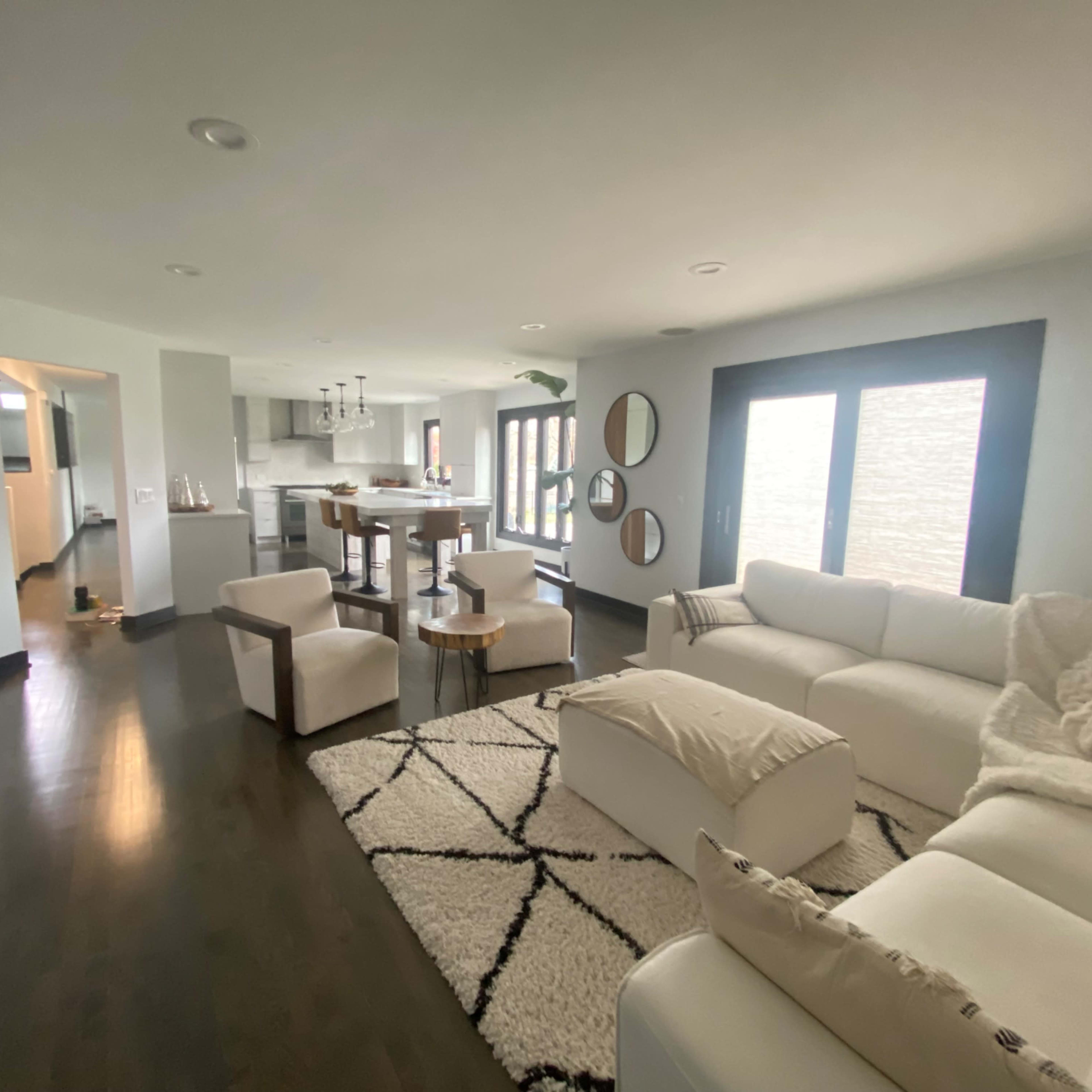 A modern, open-concept living room with a white sectional sofa, a round wooden coffee table, and large windows letting in natural light.