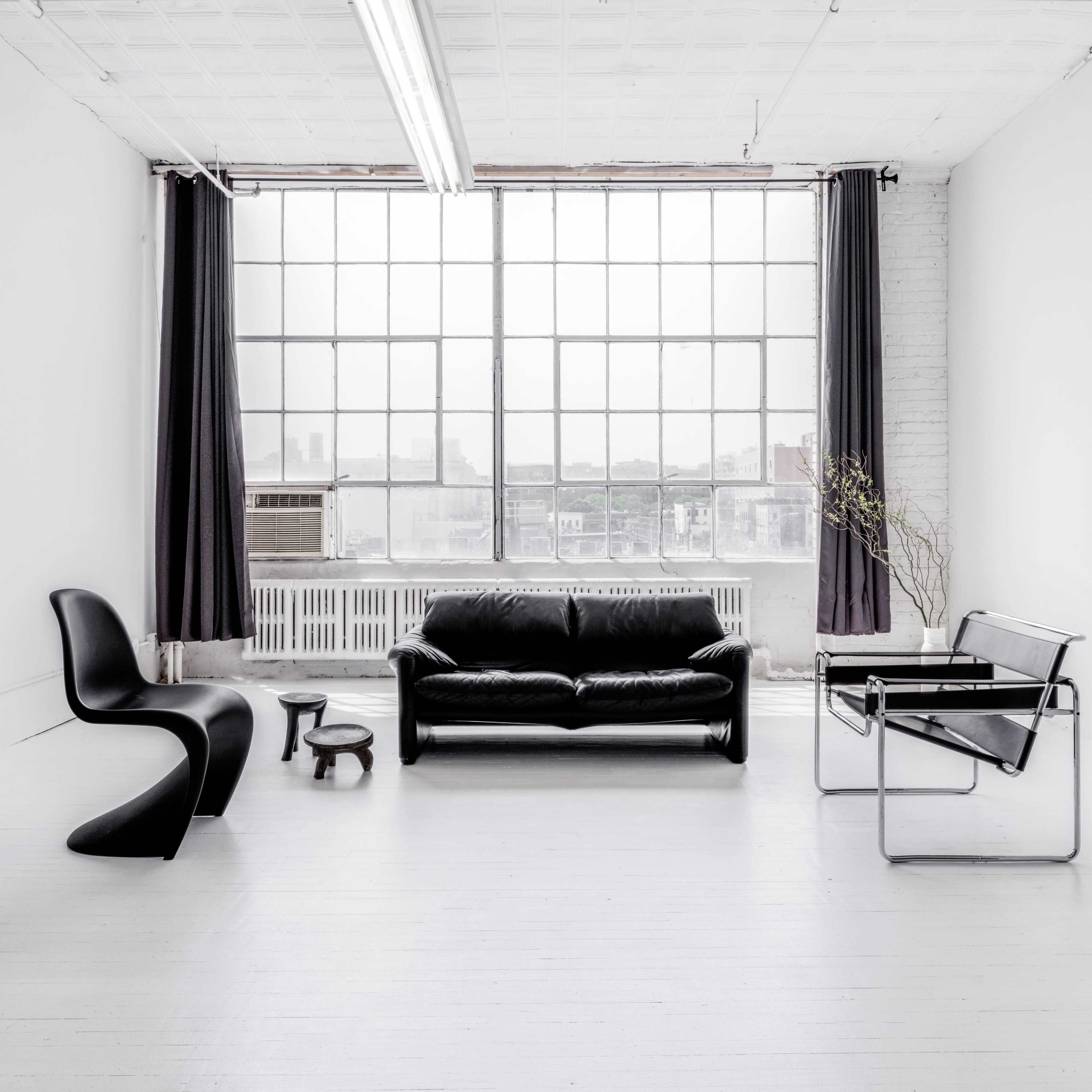 A minimalist room features a black leather sofa, a modern chair, a small table, and a large window with sheer curtains.