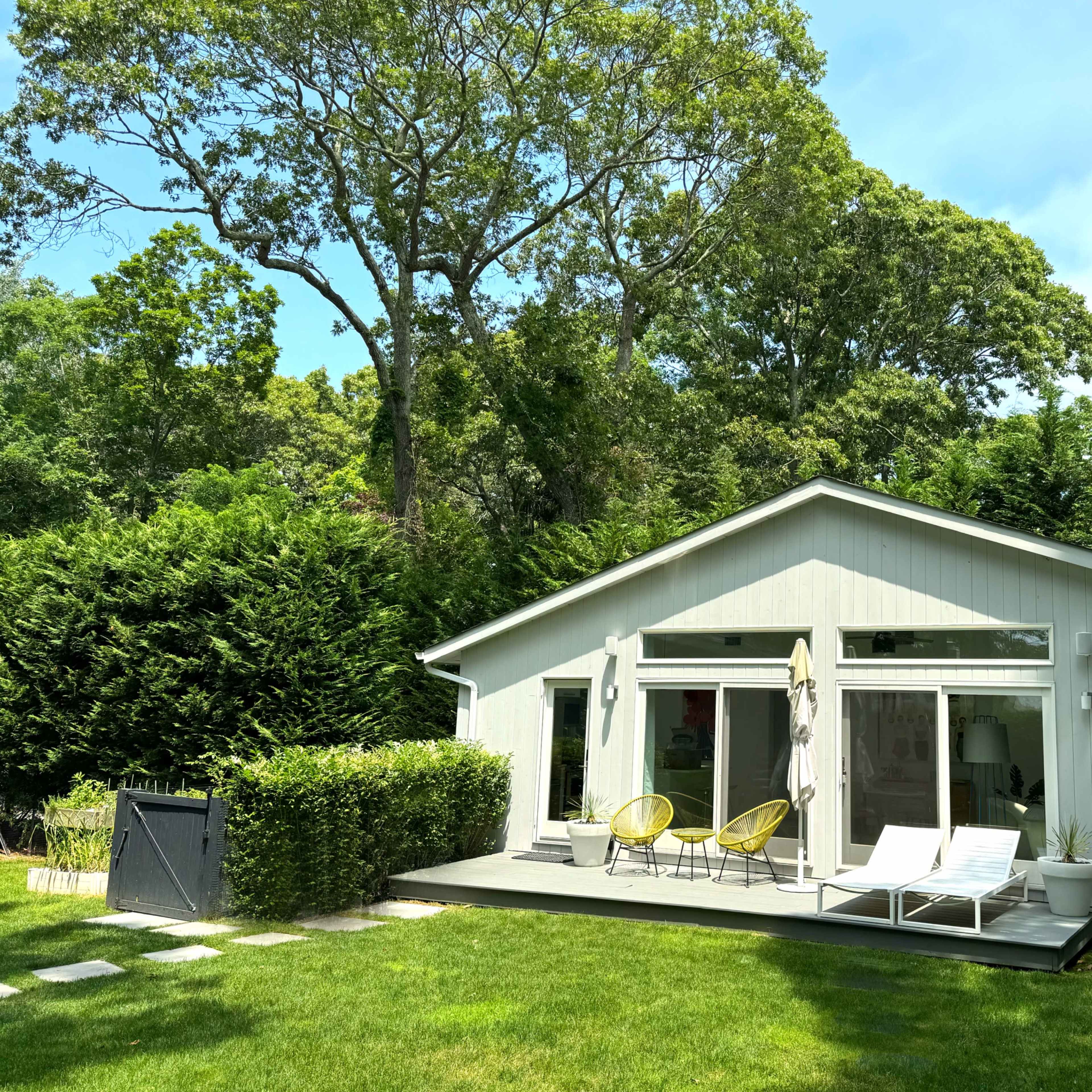 A modern single-story house features large windows and a deck with lounge chairs, surrounded by lush greenery.