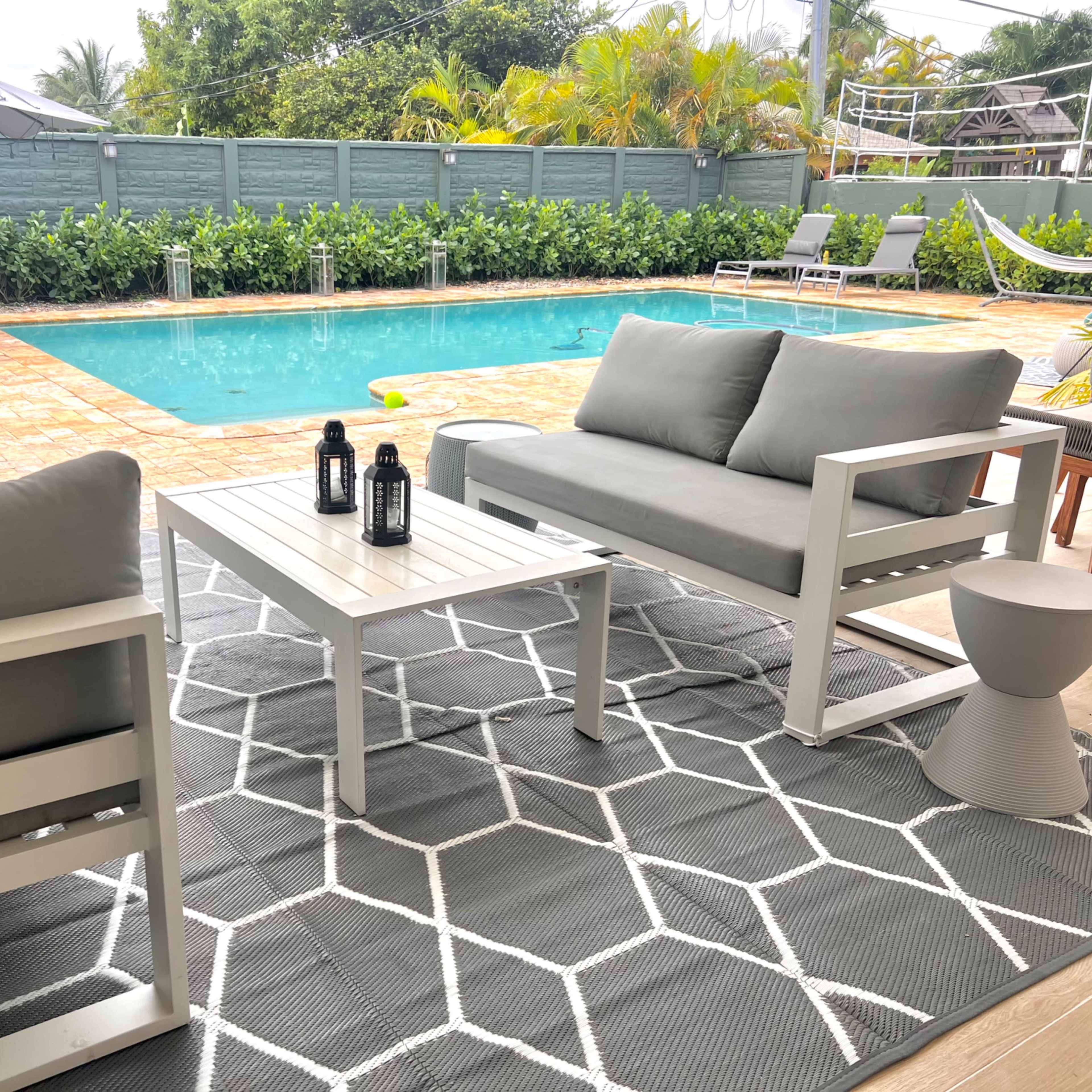 The scene features a patio area with two light gray couches and a small table, overlooking a swimming pool surrounded by lush greenery and lounge chairs.