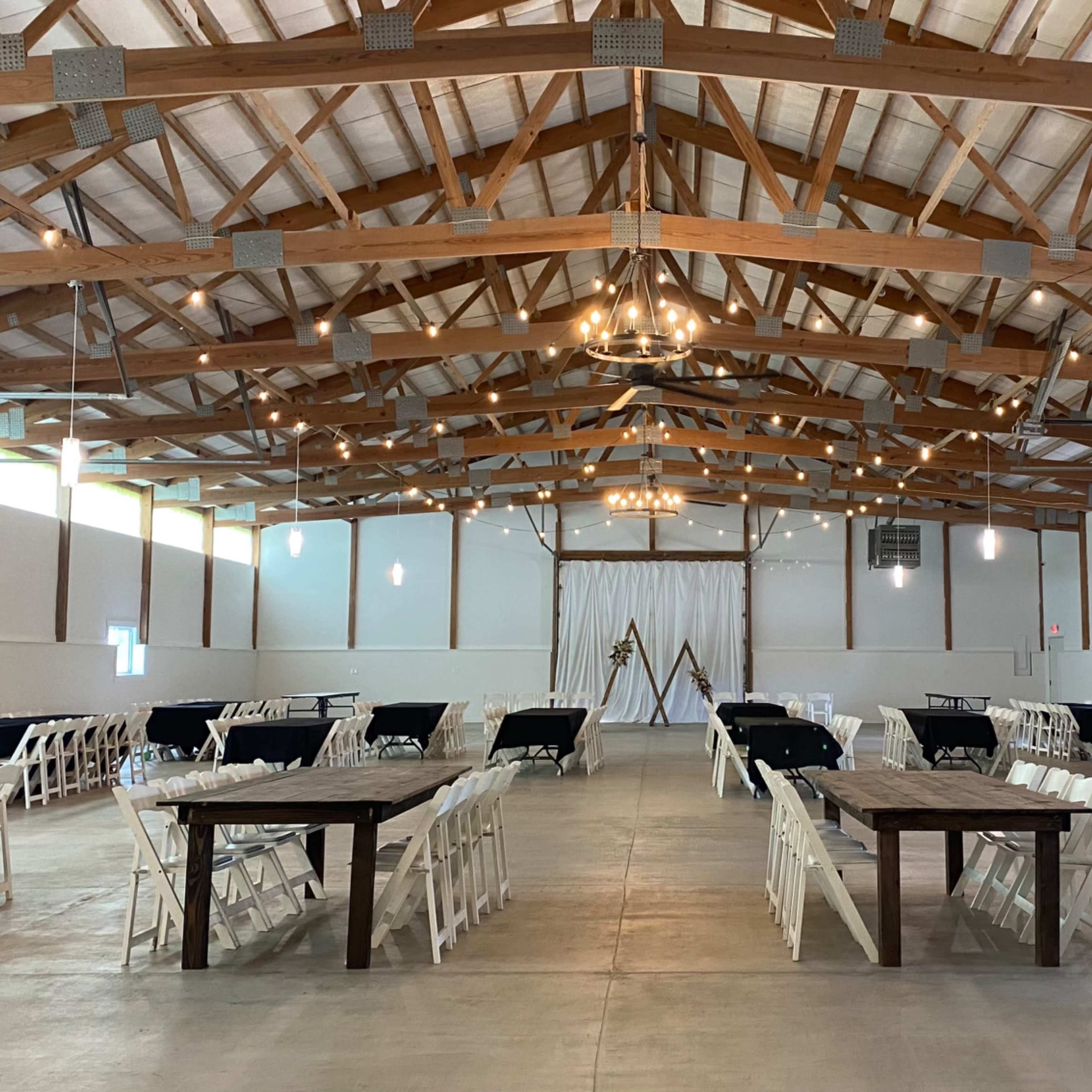 A spacious event hall with wooden beams, white folding chairs arranged around long tables, and decorative lighting.
