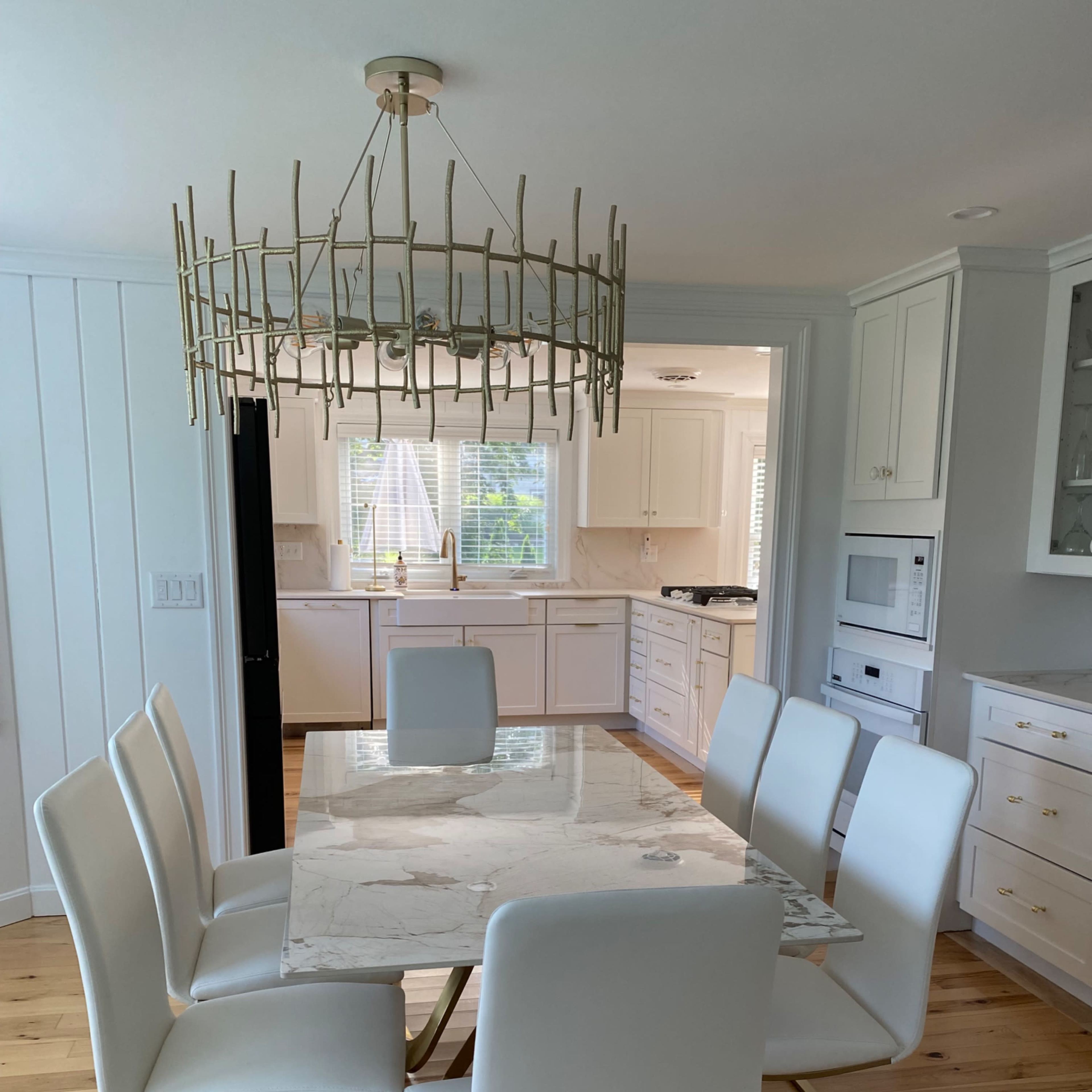 A modern dining area features a marble-topped table surrounded by white chairs, with a decorative chandelier overhead and a view of a kitchen in the background.