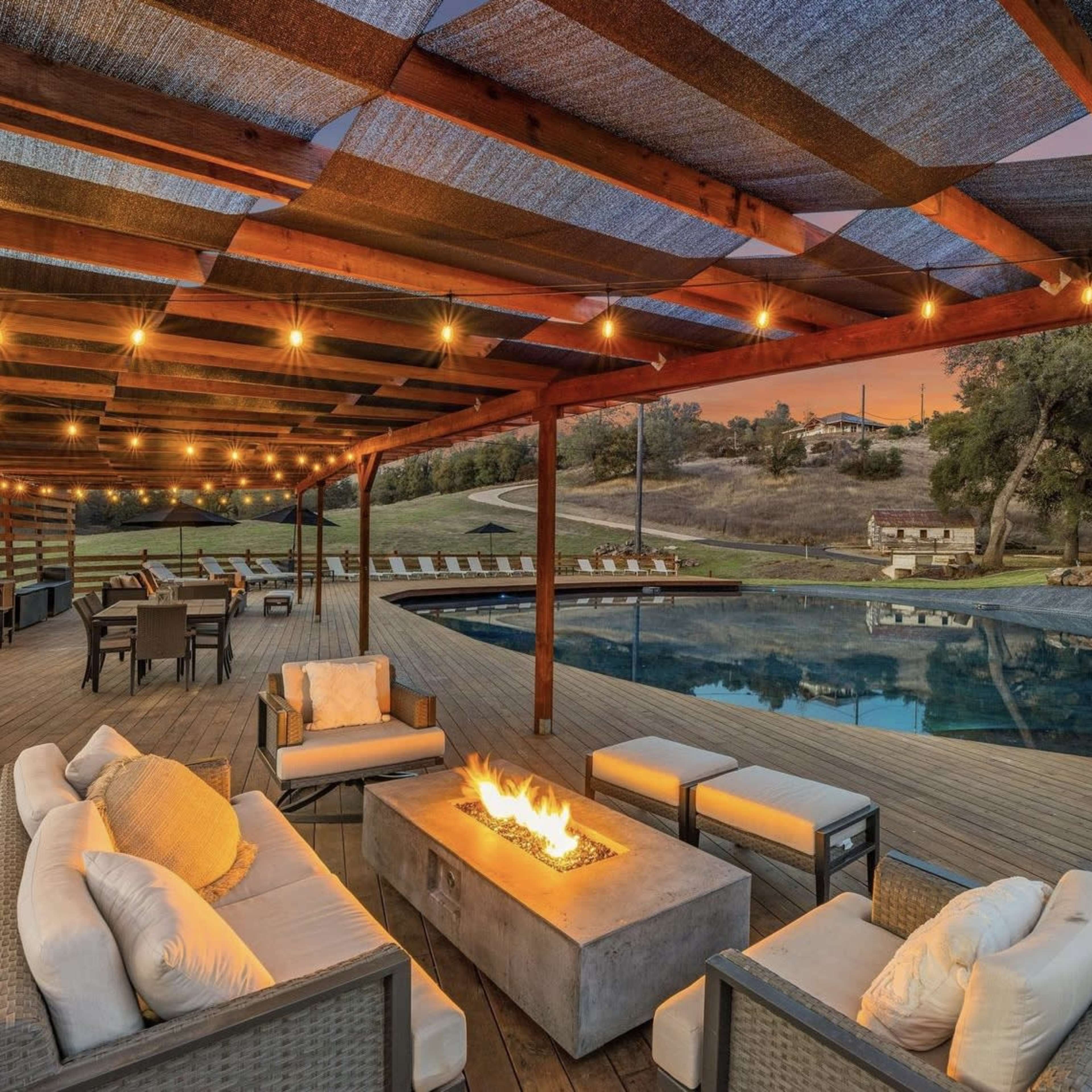 A modern outdoor patio features a fireplace surrounded by white sofas, with a pool and lounge chairs in the background under a wooden pergola with string lights.