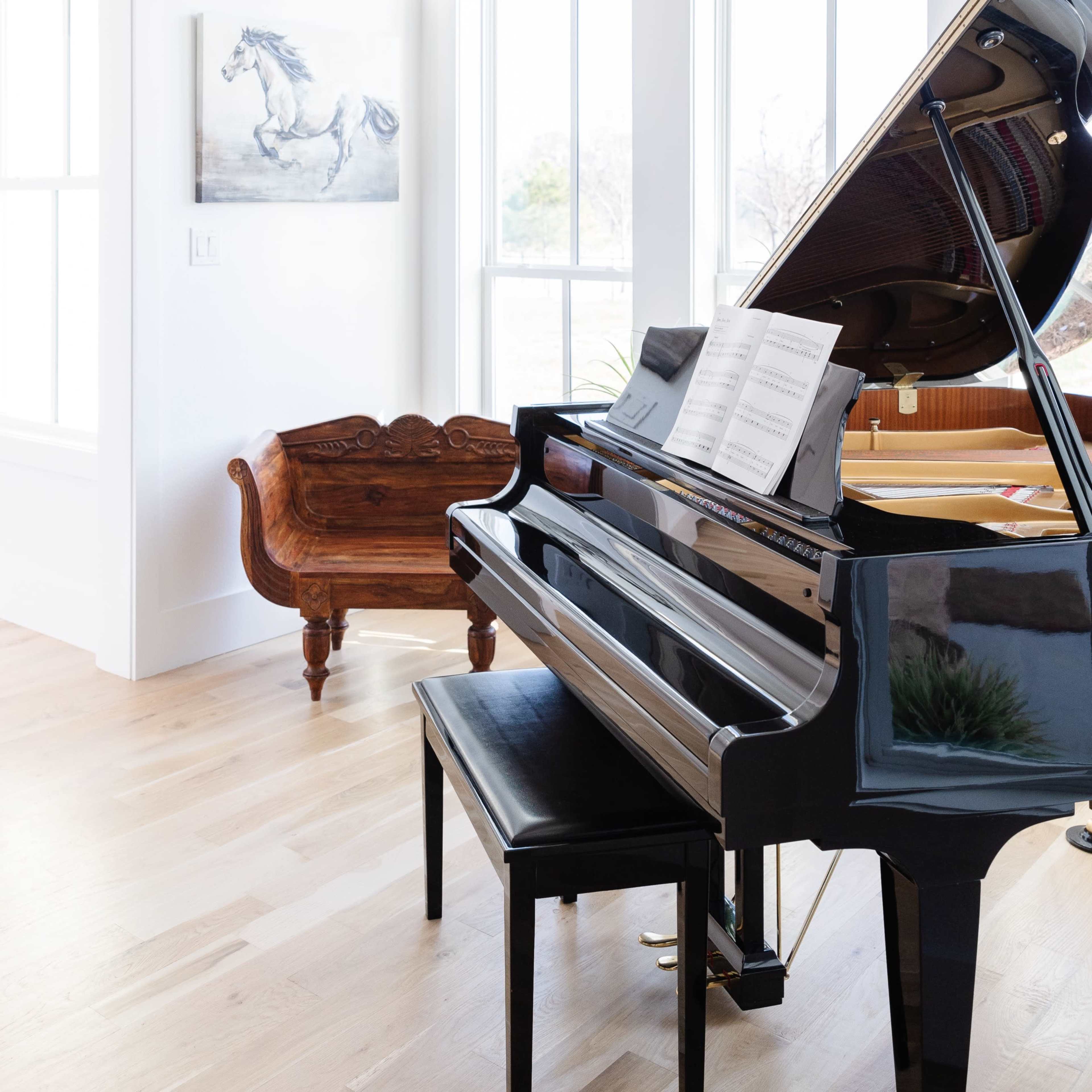 A black piano with sheet music sits beside a wooden bench in a bright room featuring large windows and a decorative light fixture.