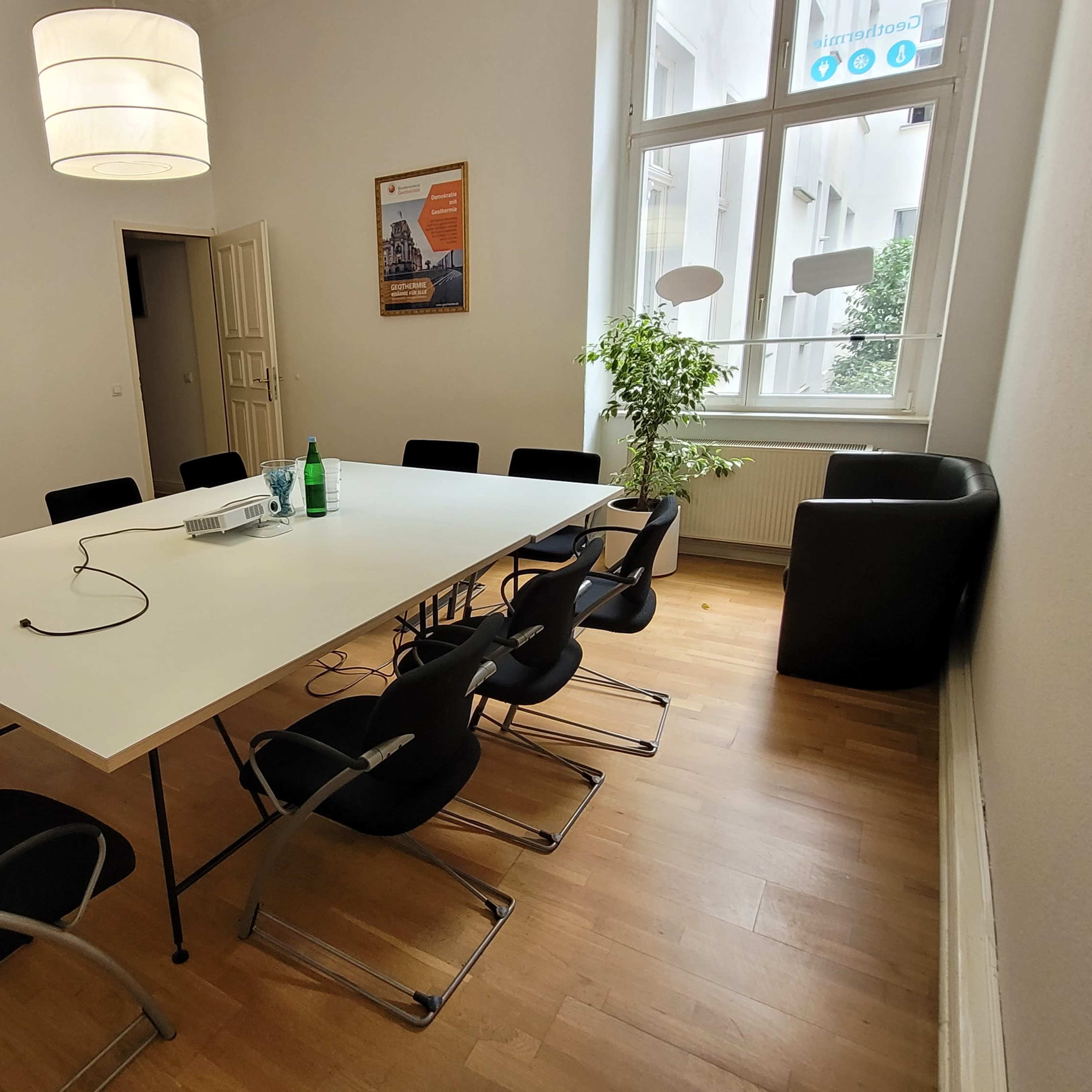 A conference room features a large white table surrounded by black chairs, with a projector on the table and a plant in the corner.