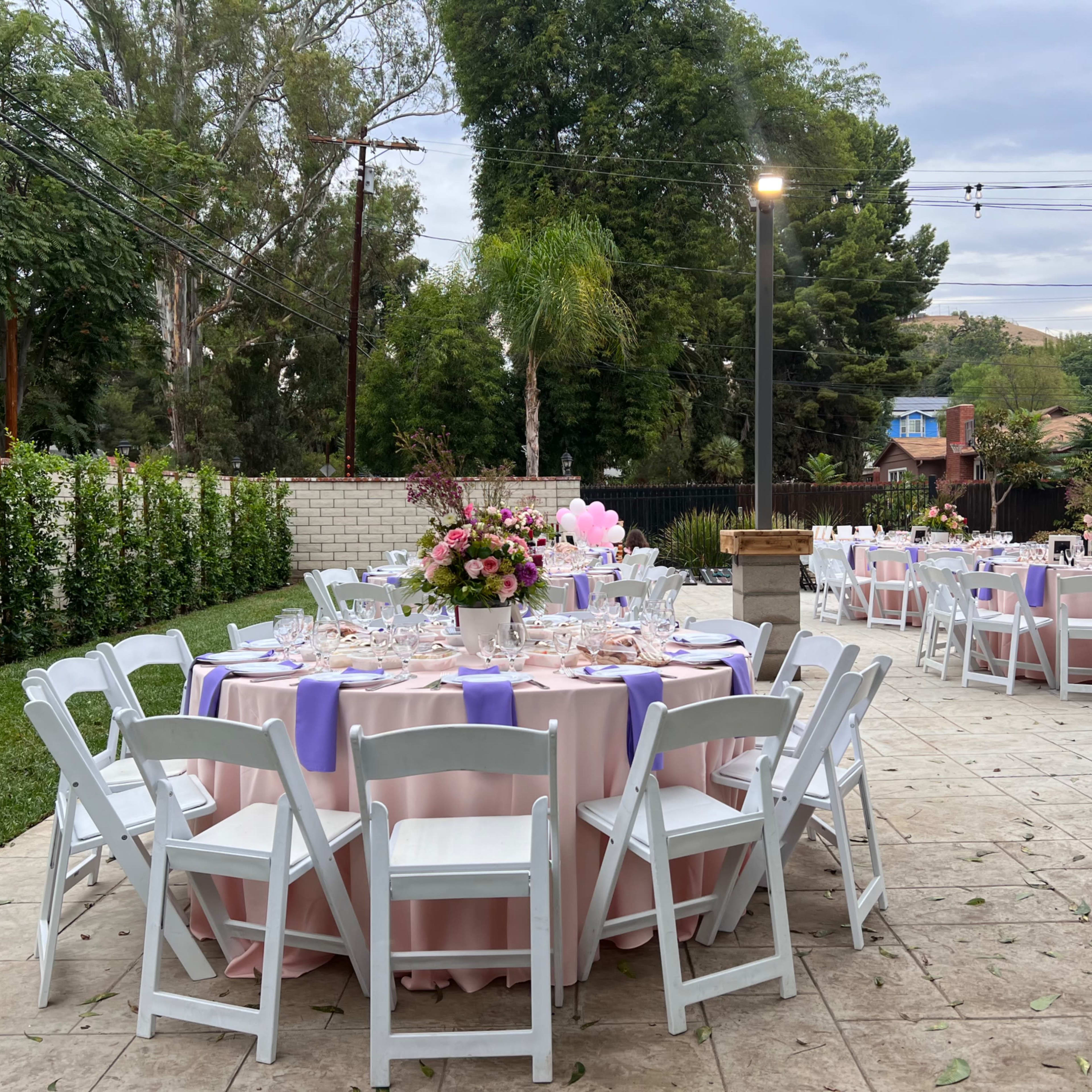 The scene features an outdoor dining area with round tables set for a formal event, complete with pink tablecloths and floral centerpieces.