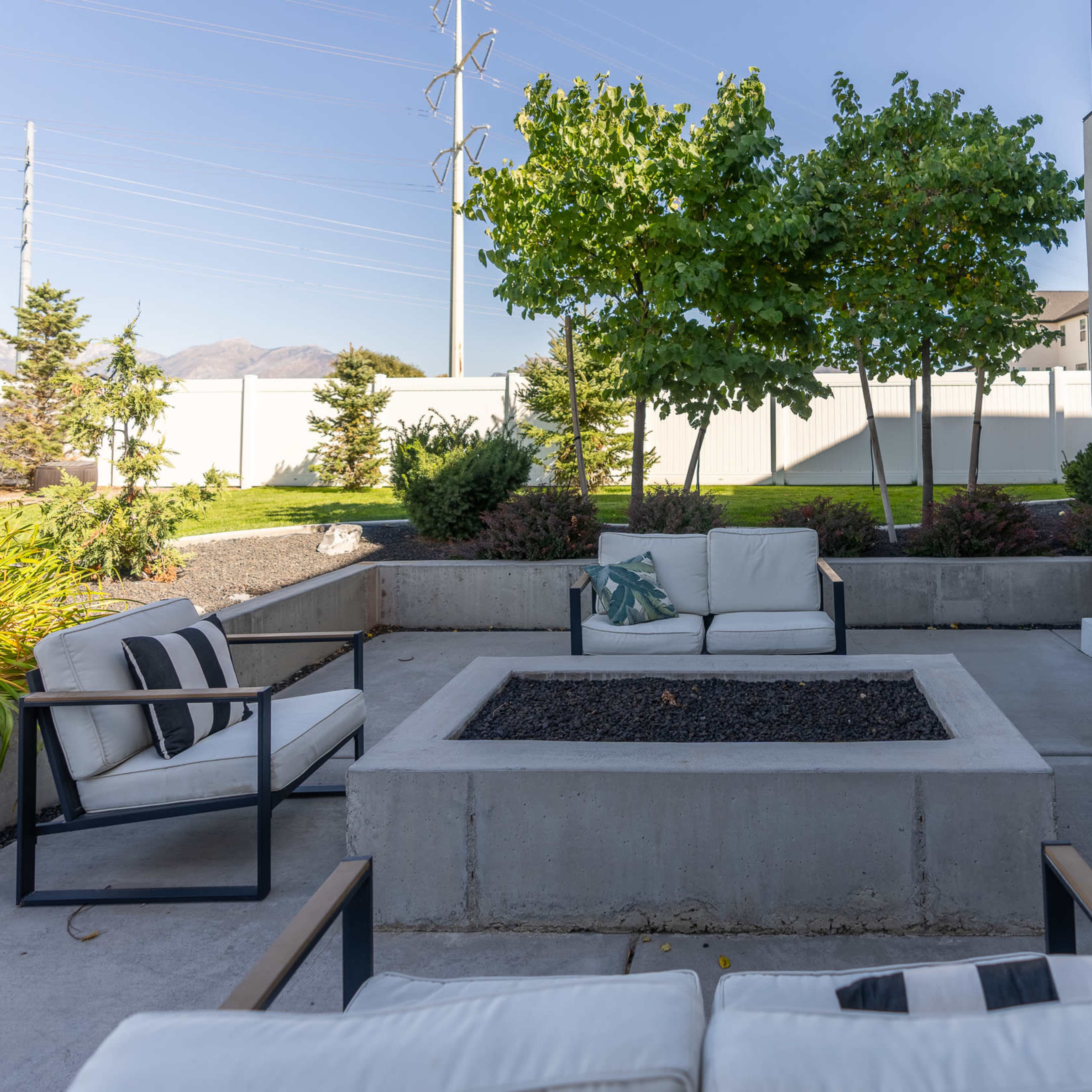 A backyard seating area with four chairs arranged around a rectangular fire pit, surrounded by low greenery and a fence in the background.