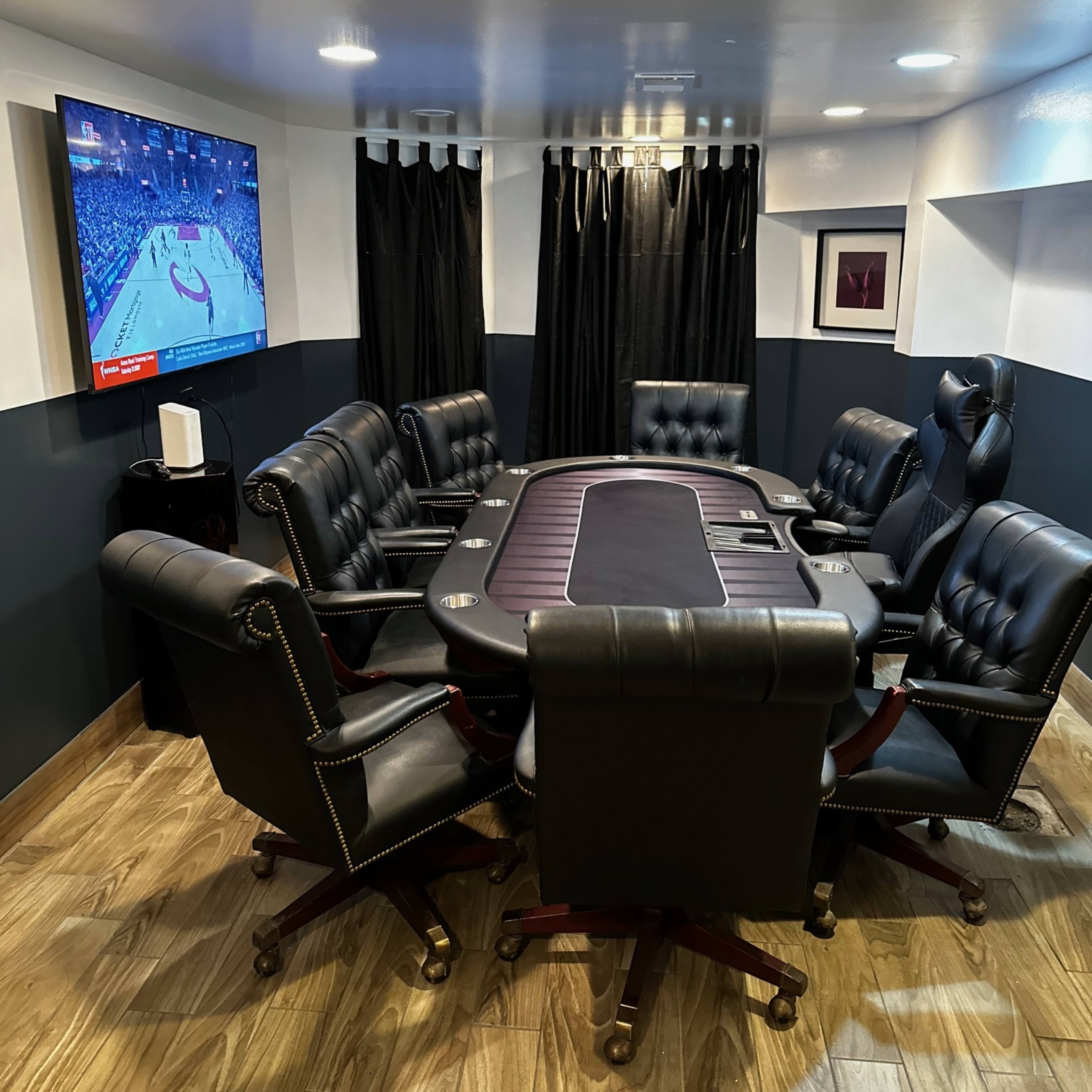 A poker room features a central table surrounded by eight black leather chairs and a television showing a sports game on the wall.