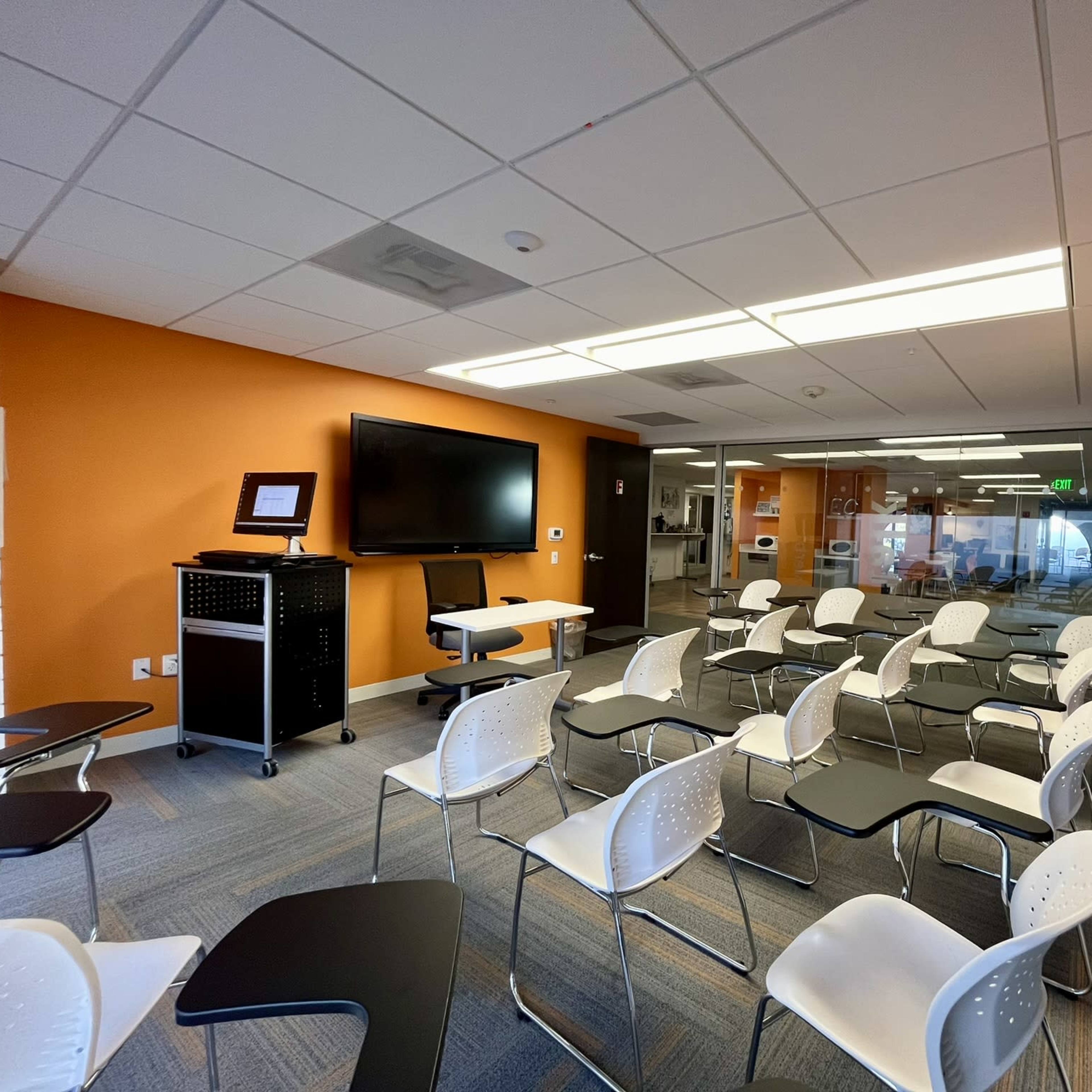 A classroom setup with rows of white chairs and a table at the front, featuring a television screen and a laptop on a cart.