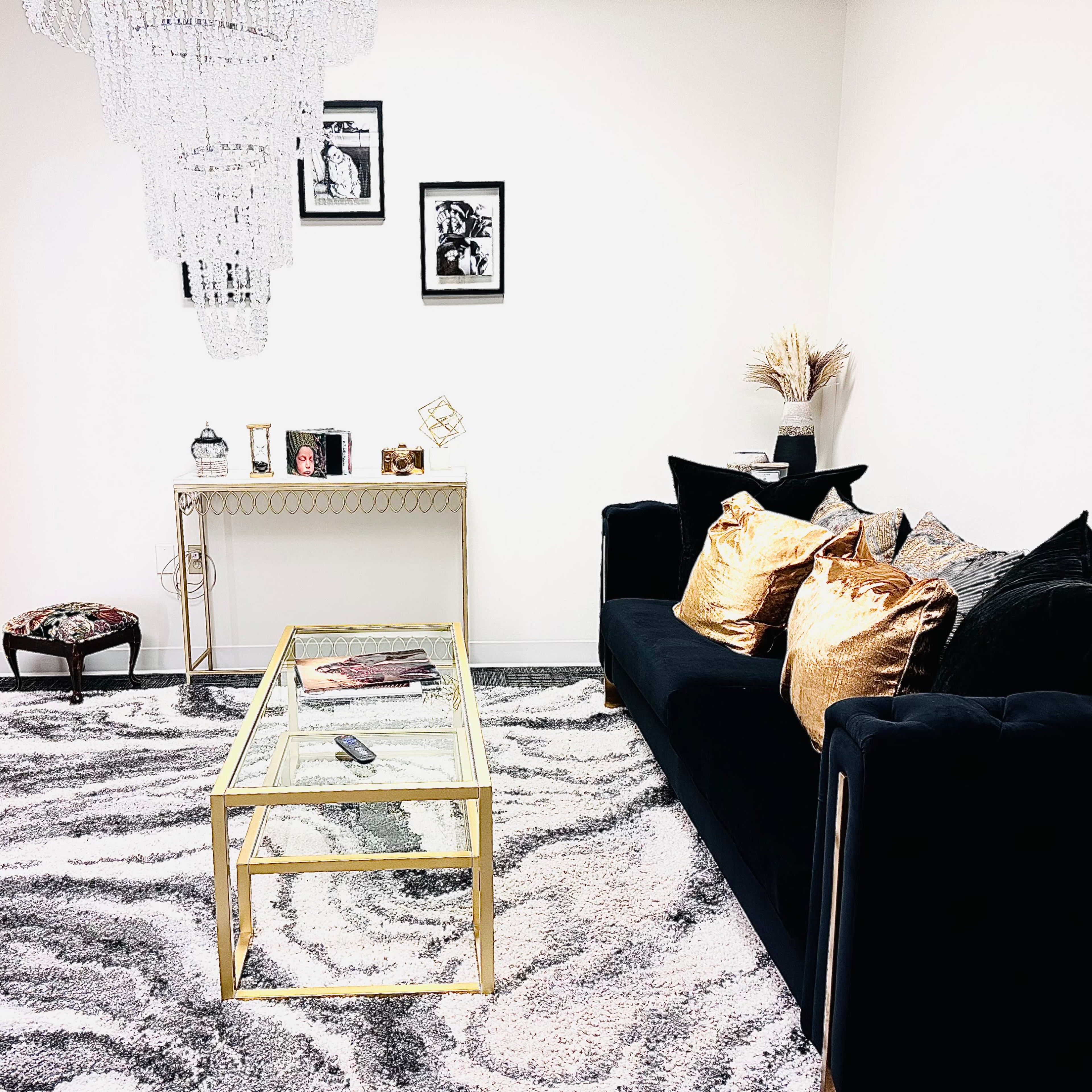 A stylish interior featuring a black sofa with decorative cushions, a gold-framed glass coffee table, a dress form, and a chandelier, all set on a patterned rug.
