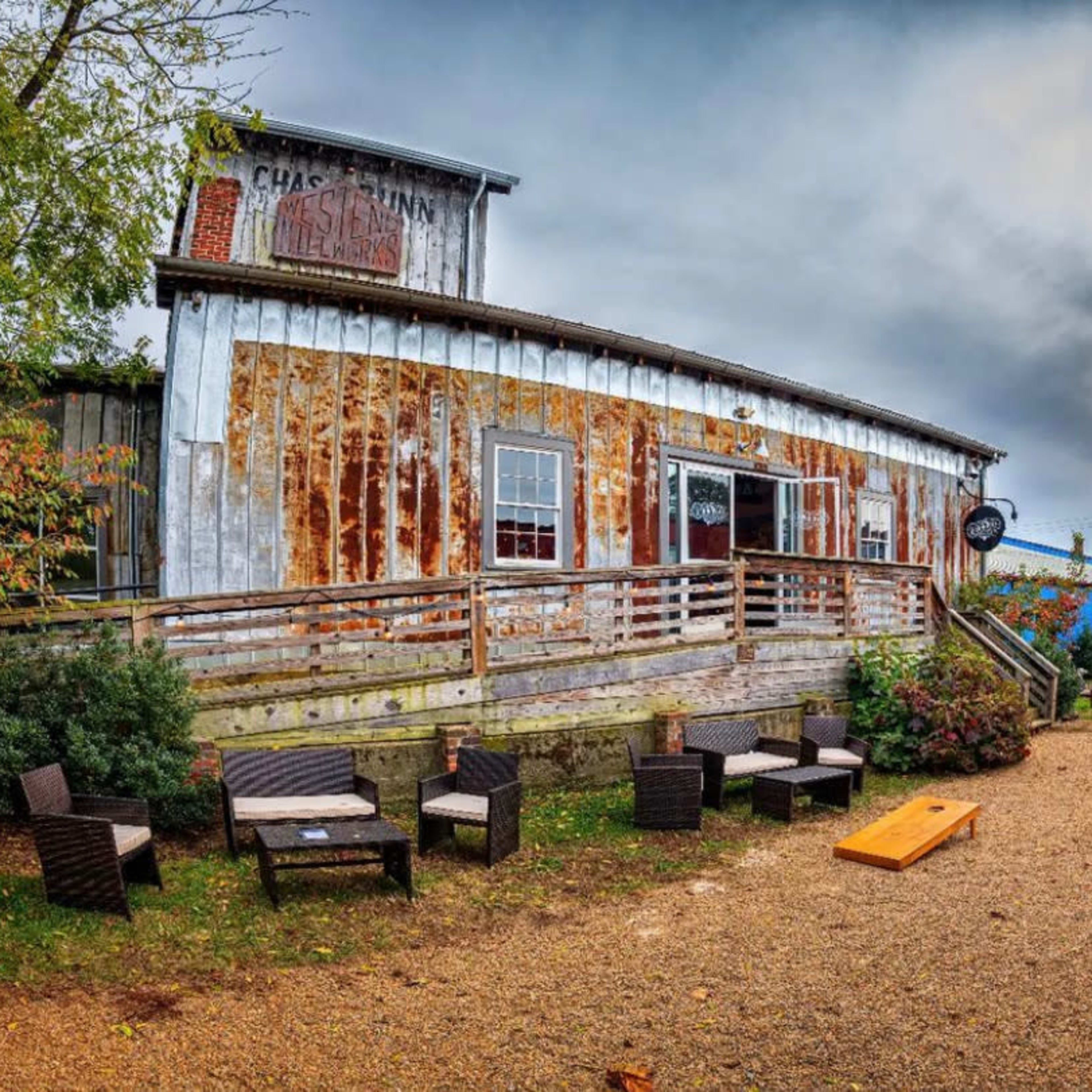 A rustic building with weathered wood siding, surrounded by a gravel path and outdoor seating areas, along with colorful canopies in the background.