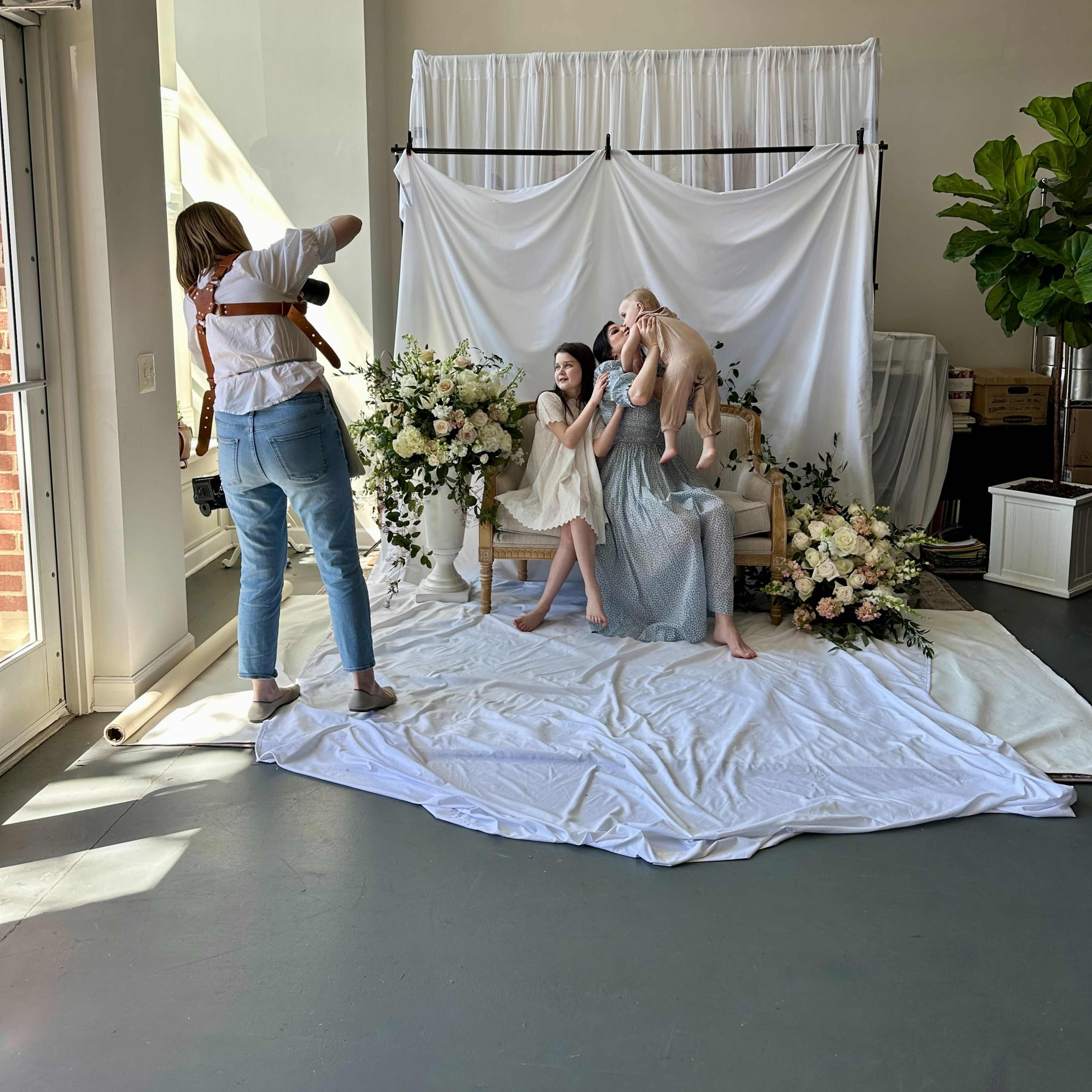 A photographer captures two seated subjects posing amidst floral decorations in a well-lit studio setting.