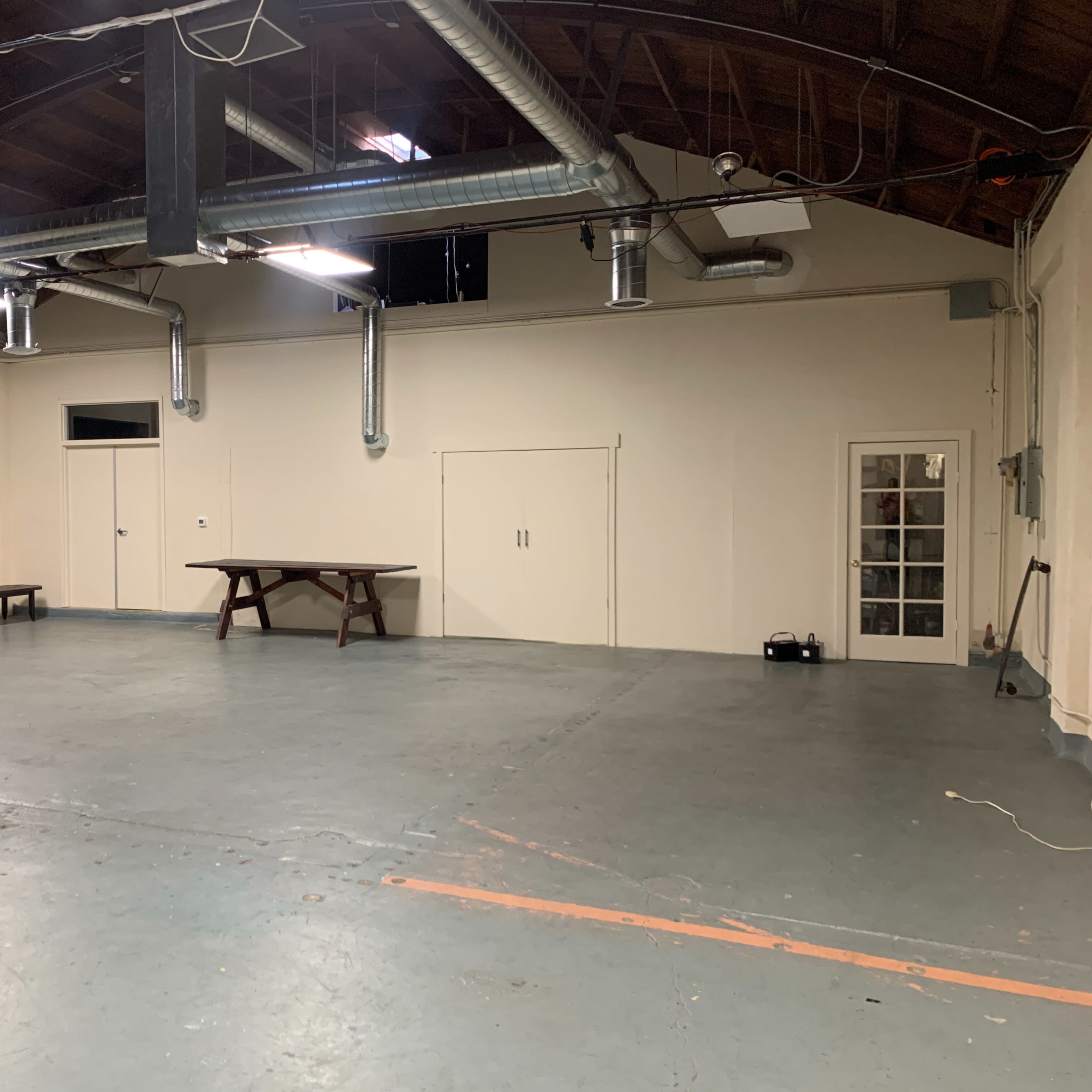 A spacious, unfurnished room with a table and a couch along one side, featuring industrial-style ventilation ducts on the ceiling.