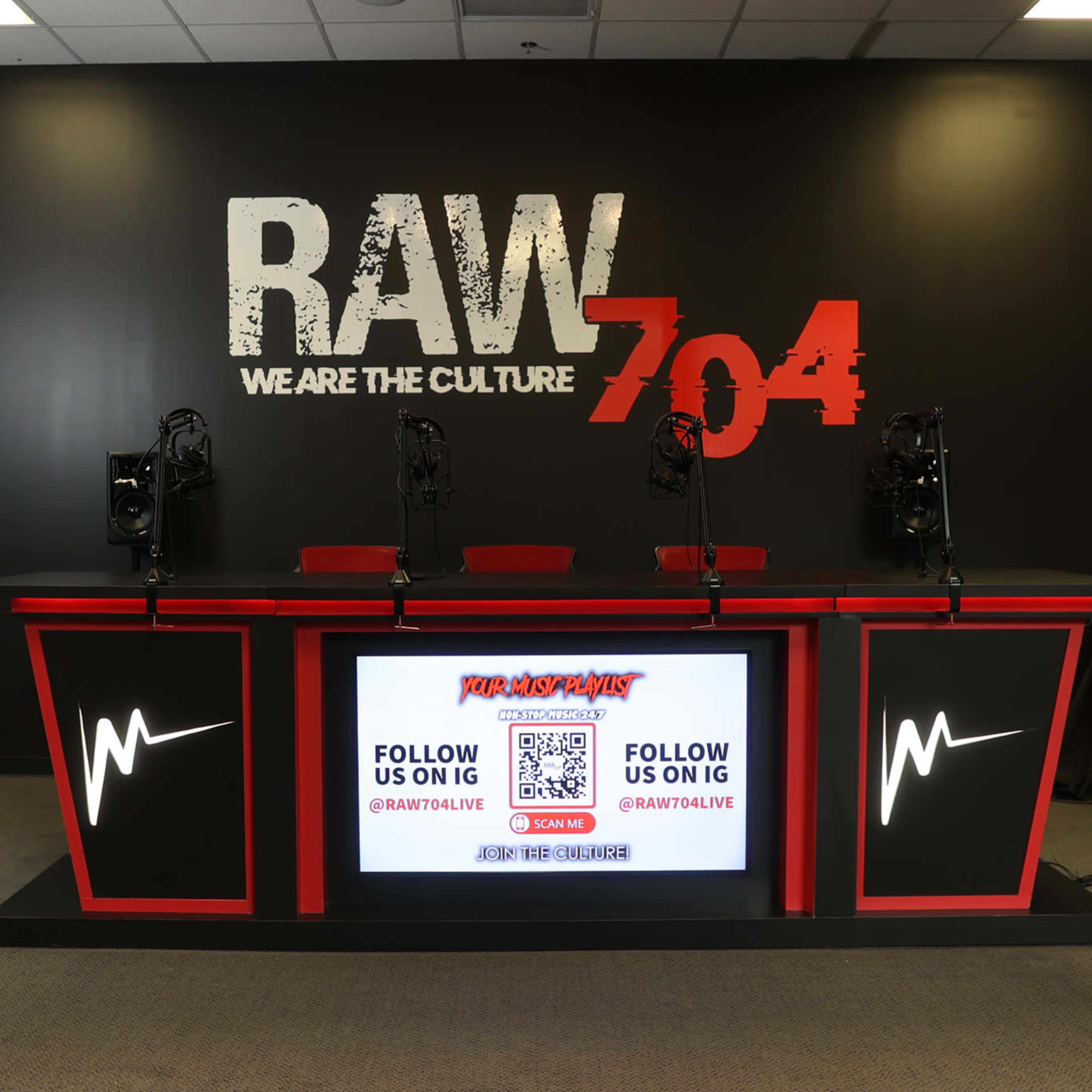 A modern broadcast studio setup with a large red and black counter, microphones, and a digital screen displaying social media handles against a black wall with the text "RAW 704."