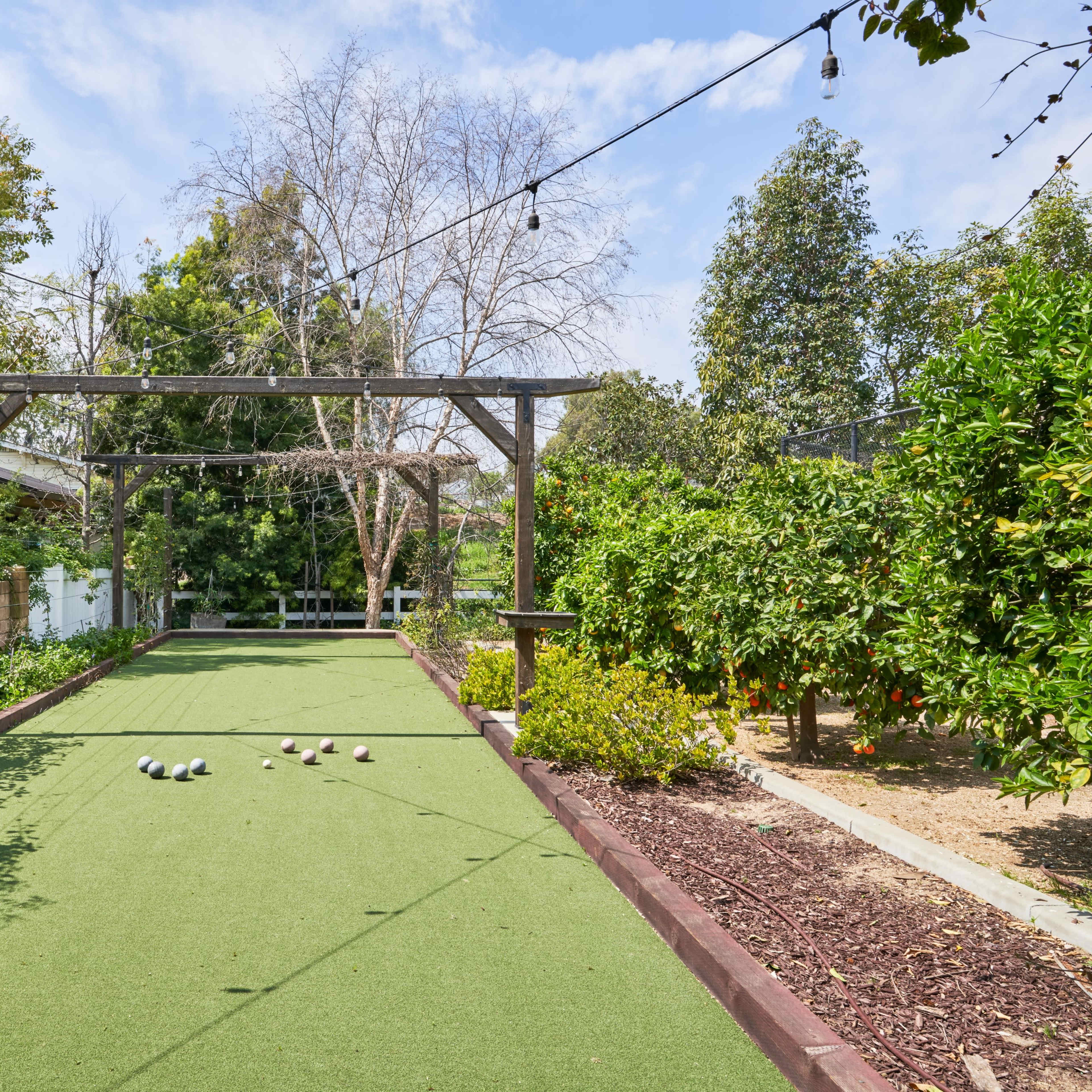 A bowling green with artificial turf surrounded by trees and plants, under a blue sky.