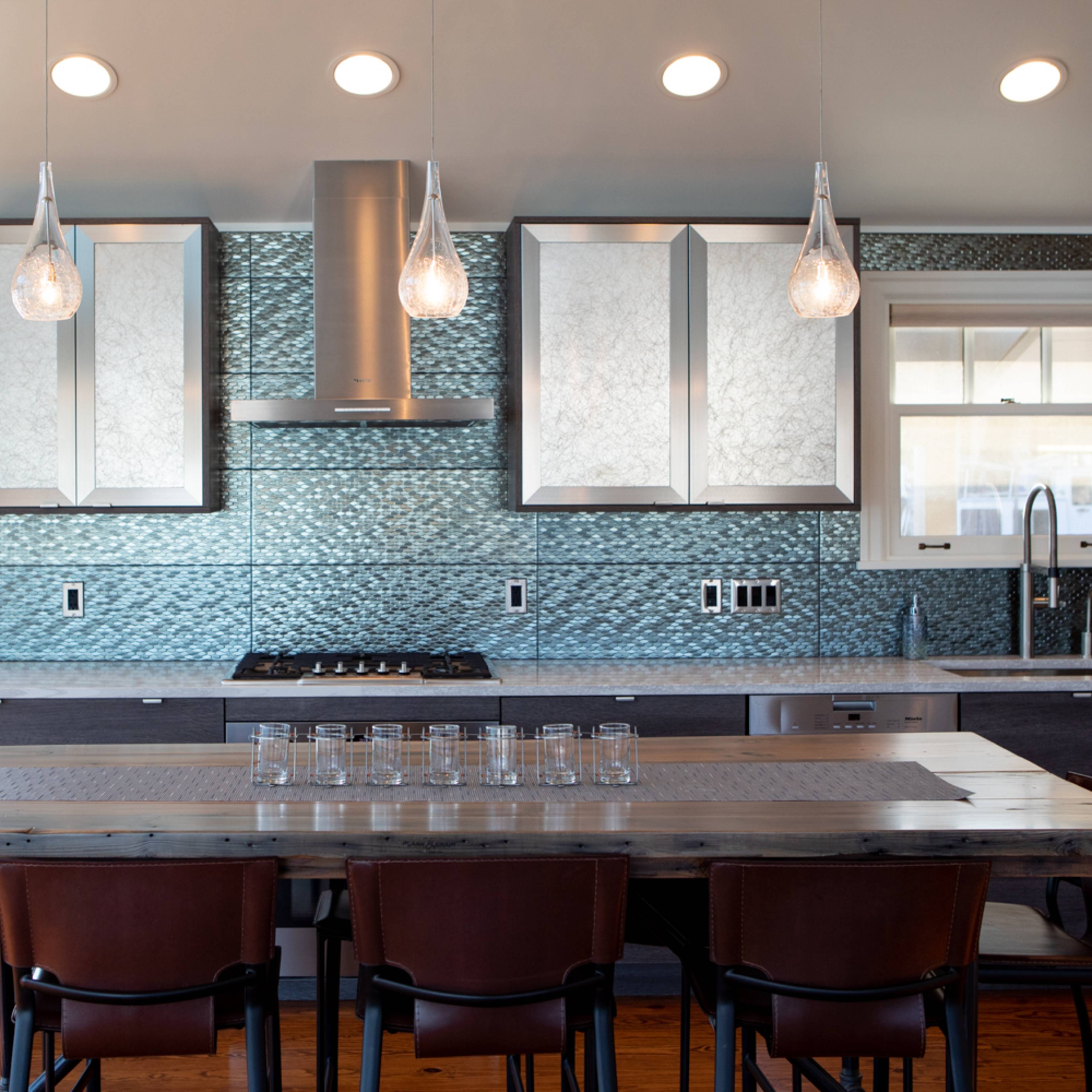 A modern kitchen features a central island, sleek cabinetry, and a glass tile backsplash, complemented by pendant lighting and a wooden dining table.