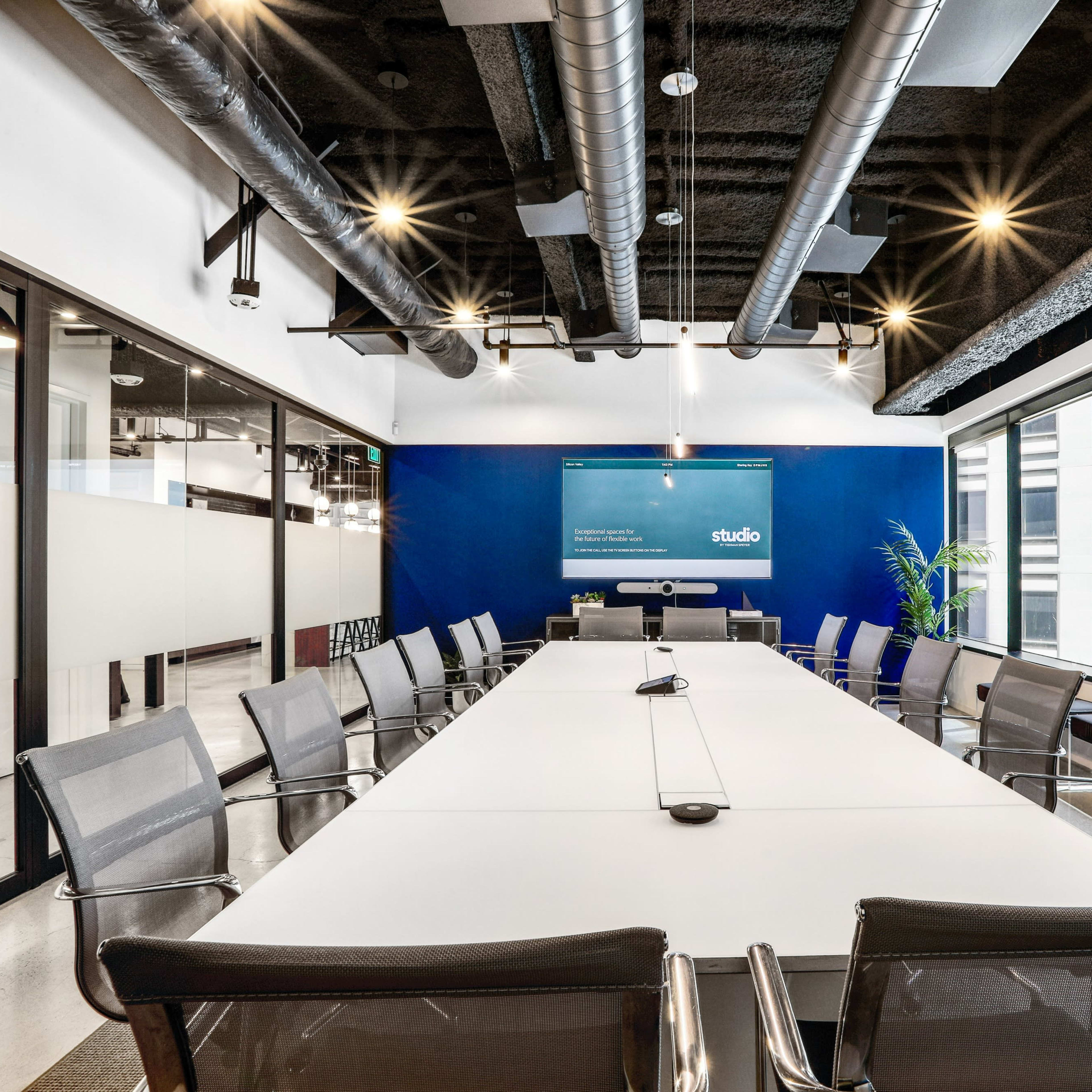 A modern conference room features a long white table surrounded by metal chairs, with large windows offering views of a cityscape.