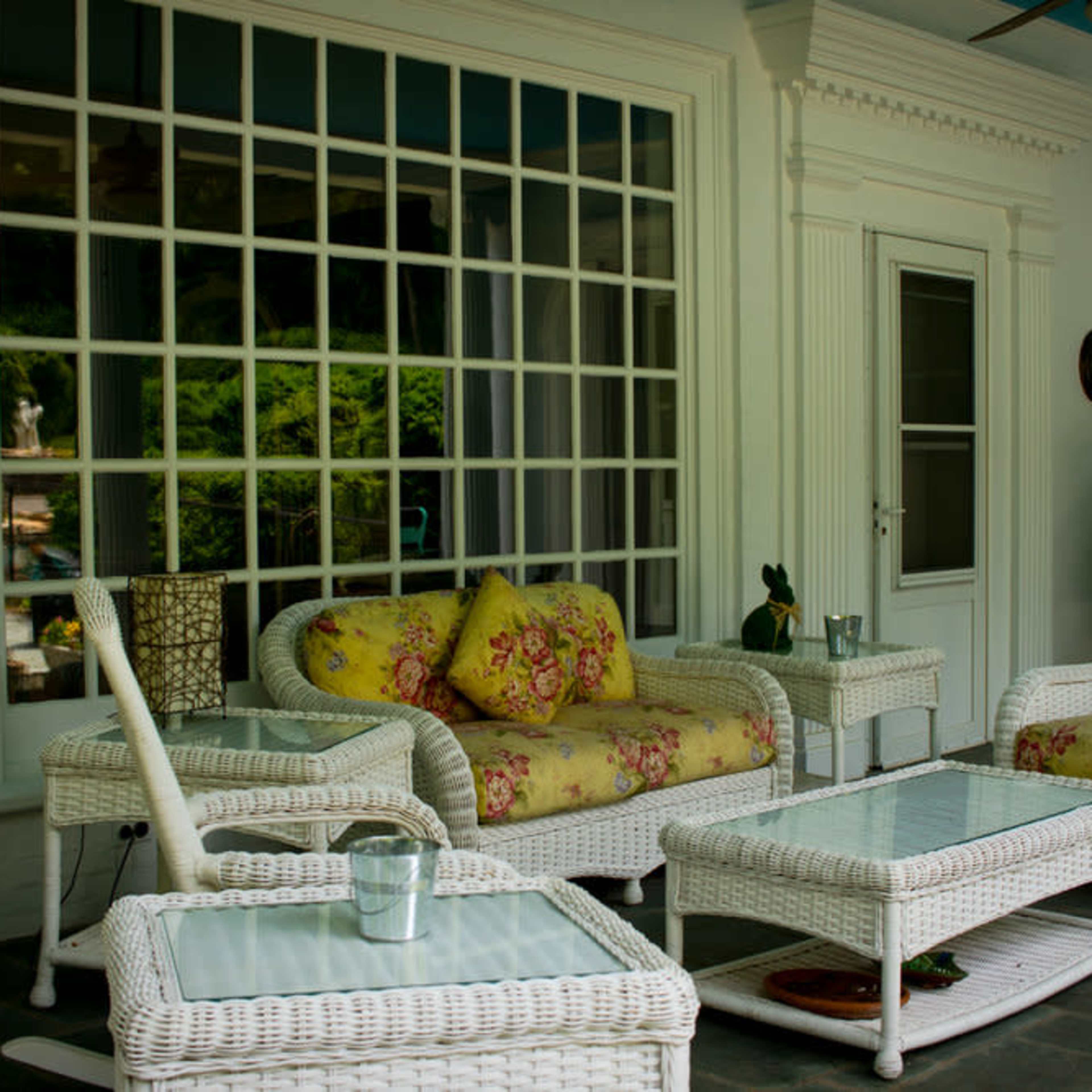 A porch area with white wicker furniture, including two chairs and a coffee table, arranged around a glass-topped table, with a large window in the background.