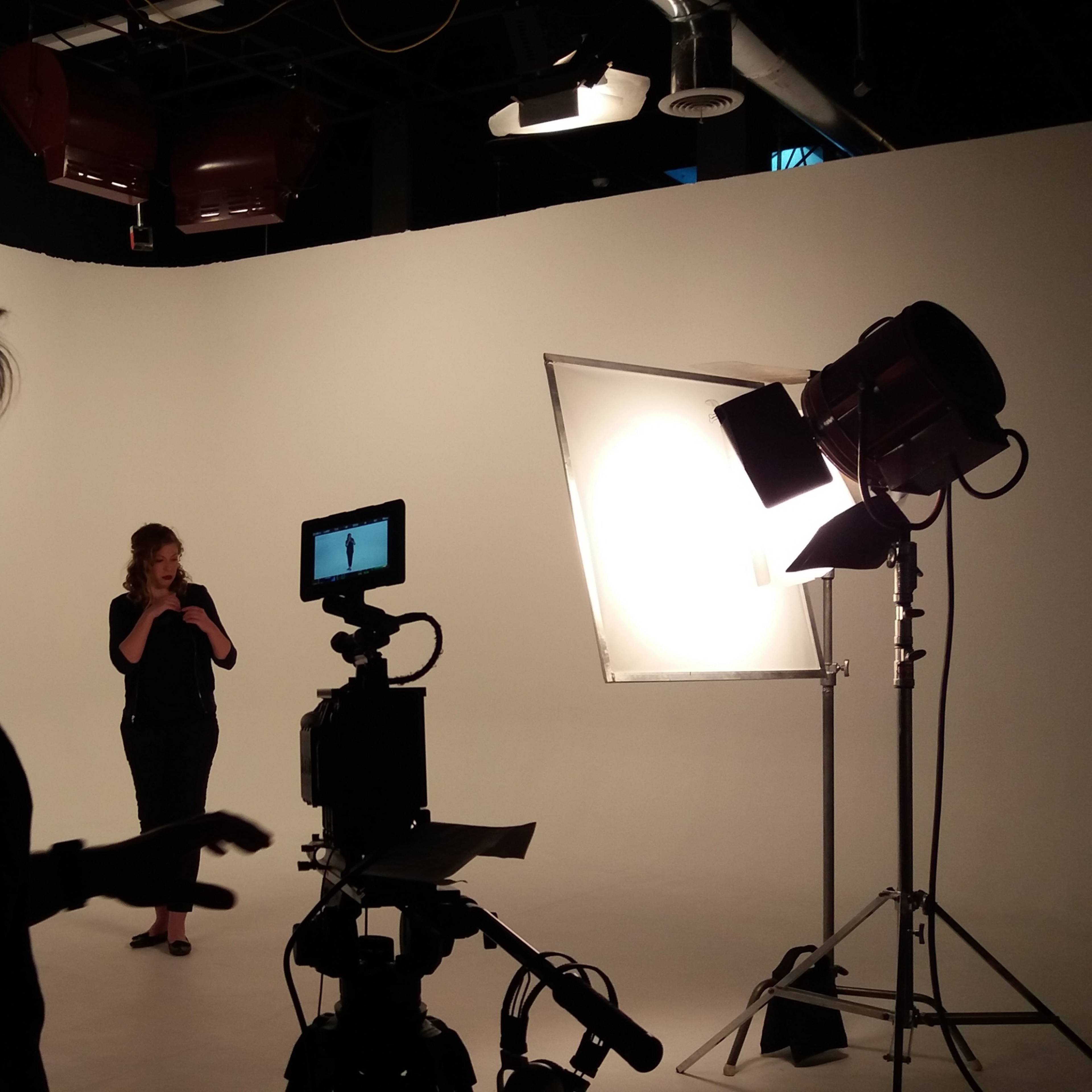 A person operates a camera while another stands in front of lighting equipment in a photography studio.