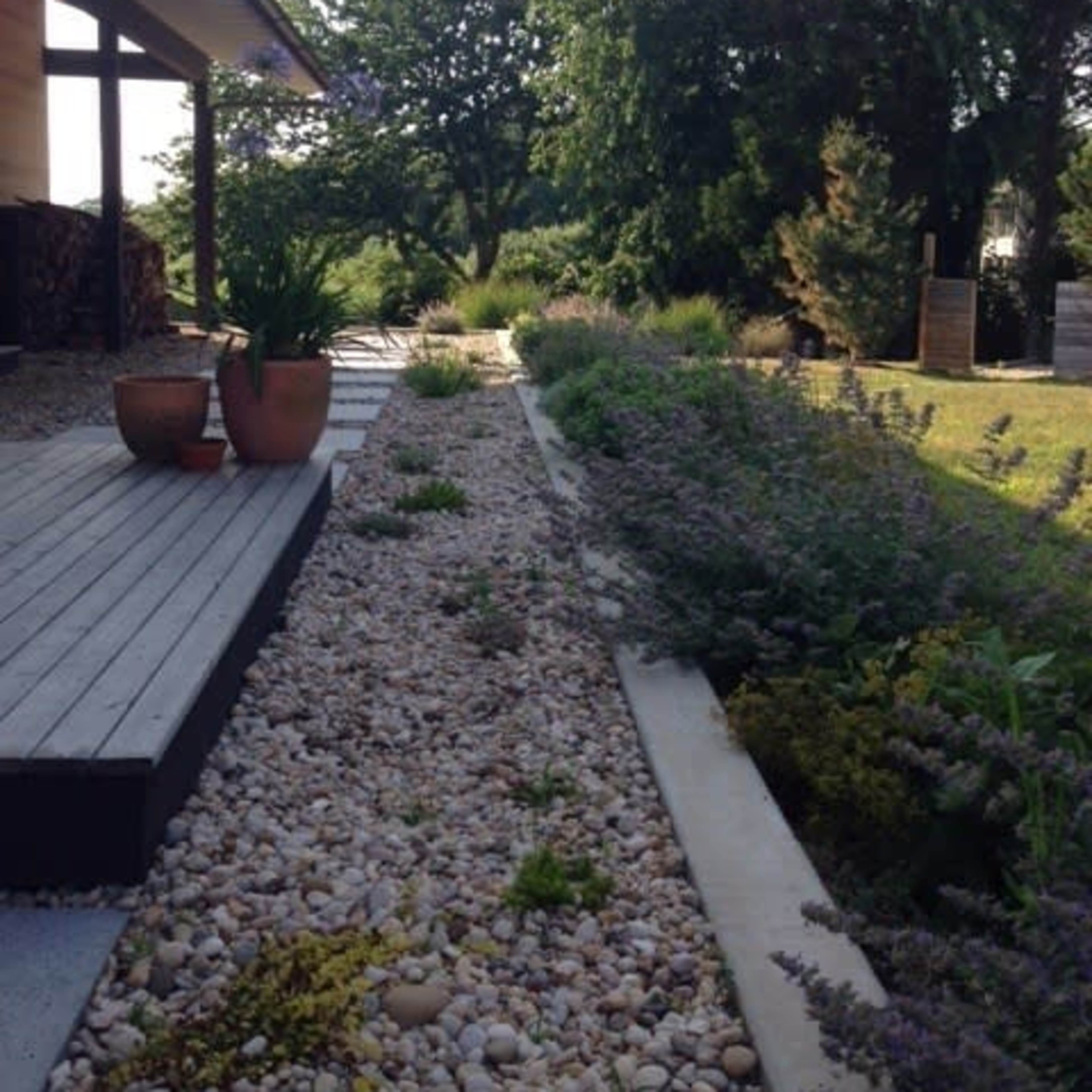 A landscaped pathway lined with stones and plants runs alongside a wooden deck, leading towards greenery in the background.