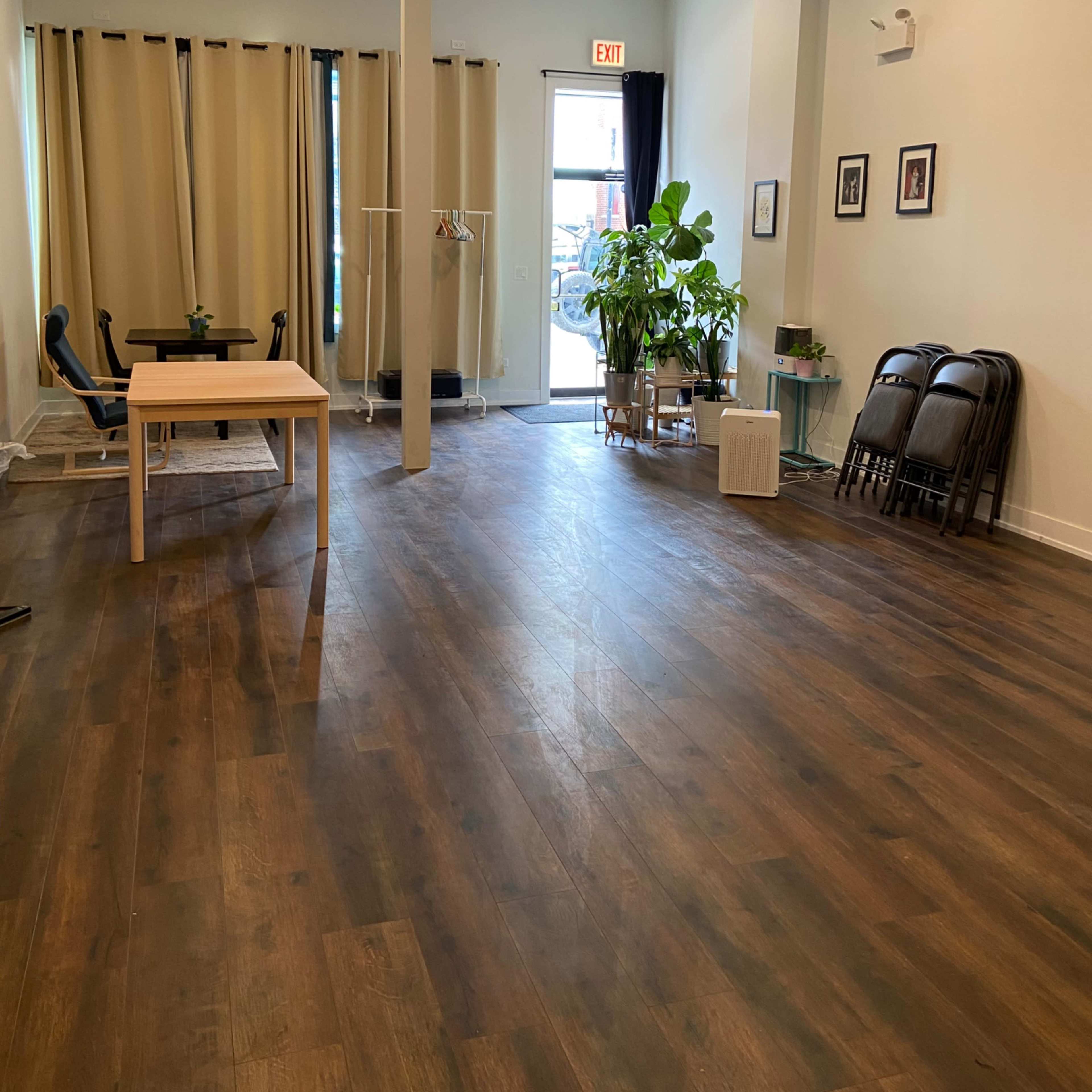 A spacious room with wooden flooring, a table, chairs, large windows with curtains, potted plants, and a door leading outside.
