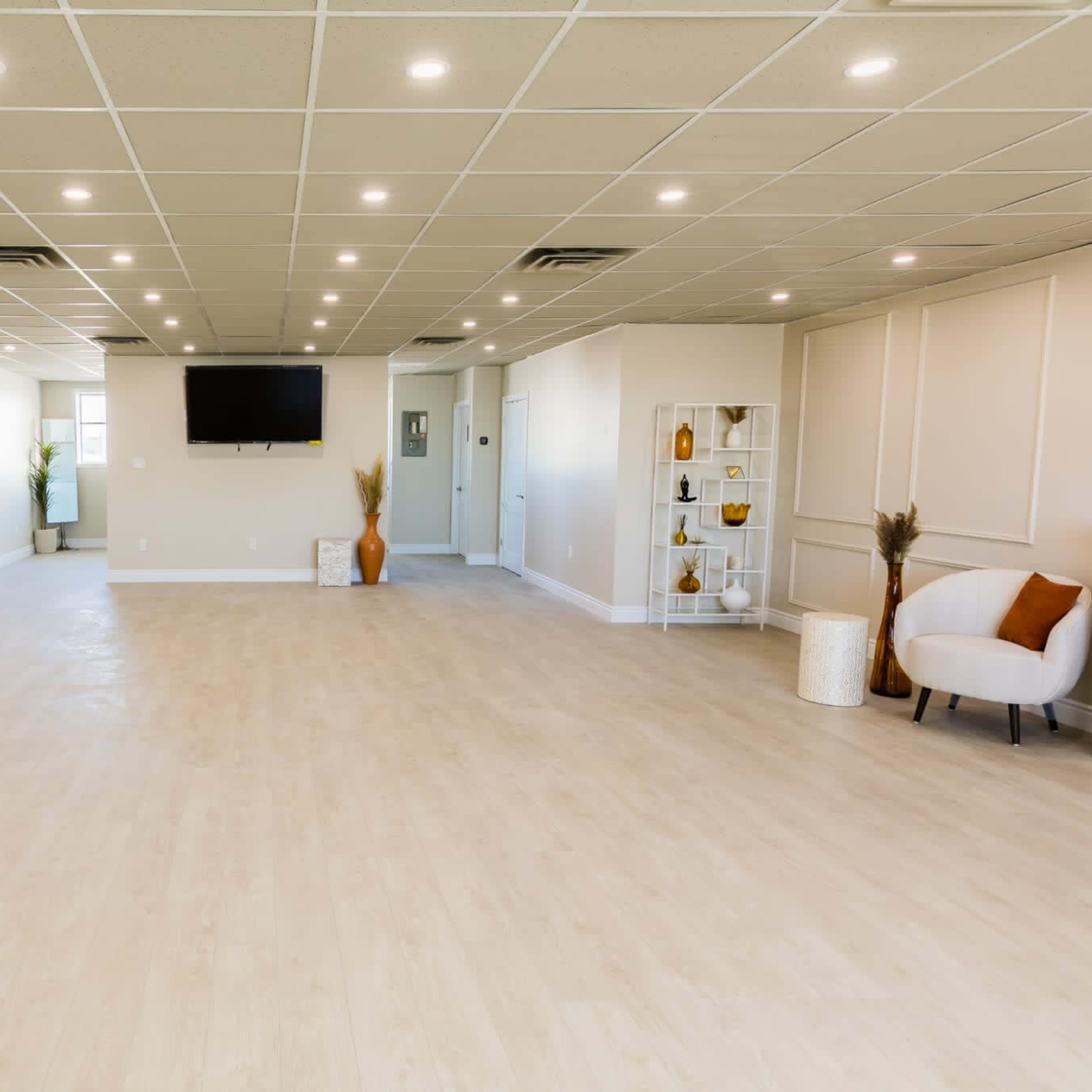 A spacious, well-lit interior room with modern decor, including a television on the wall and furnishings like a chair and decorative shelves.