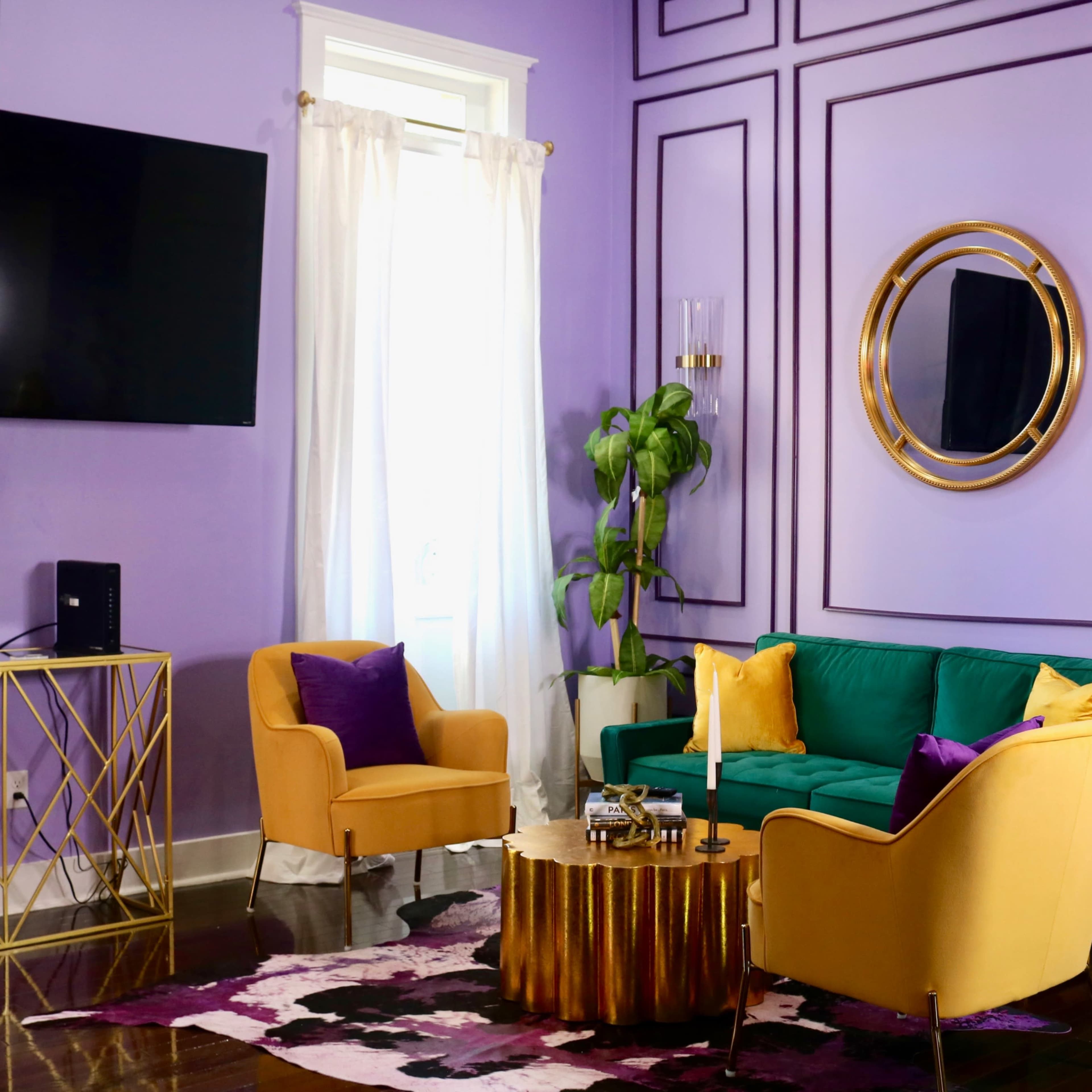 A brightly colored living room features a green sofa, yellow chairs, a round wooden coffee table, and purple walls with decorative molding.