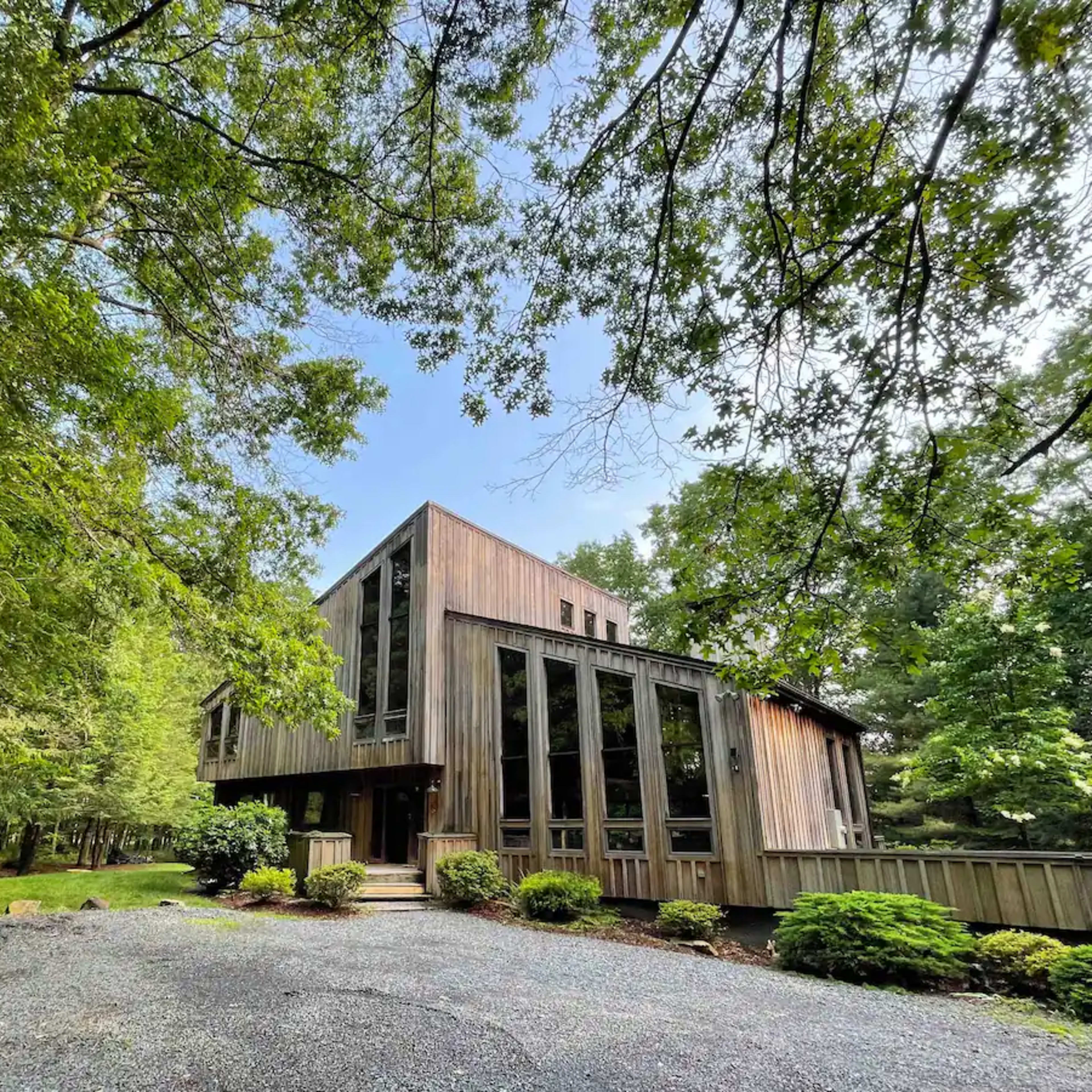 A wooden house with large glass windows is surrounded by trees and a gravel driveway.