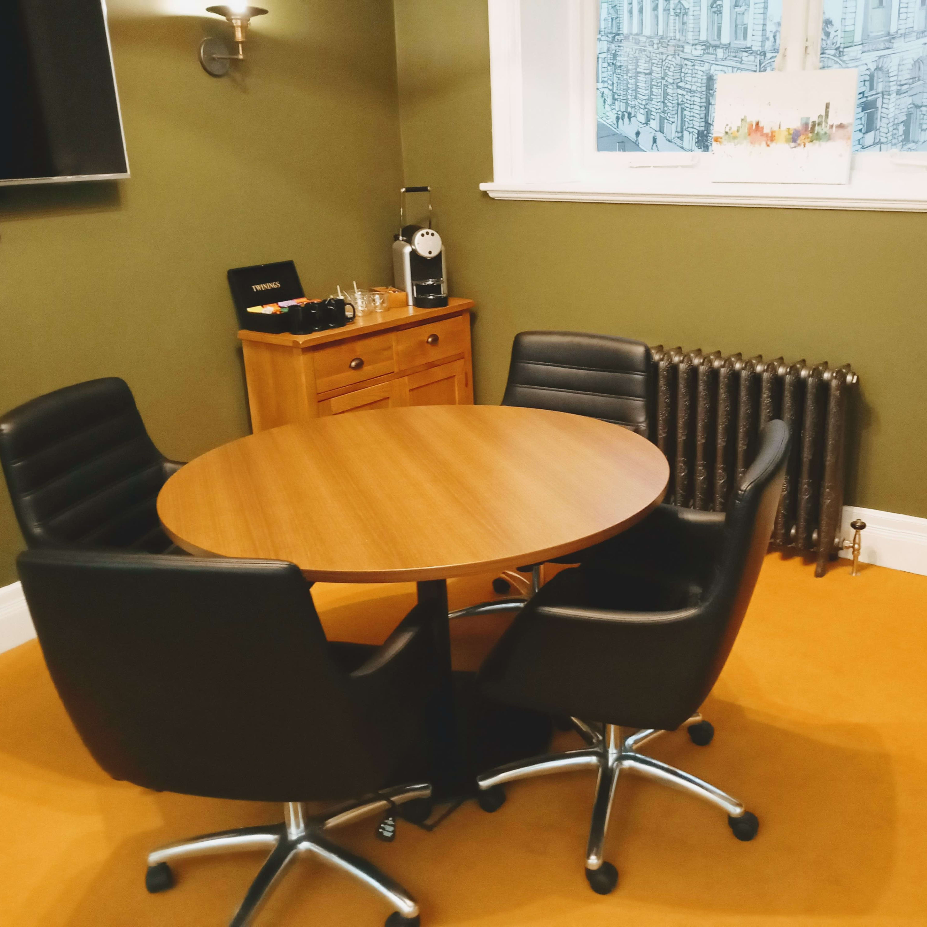 A small meeting area with a round wooden table surrounded by four black swivel chairs, a radiator, and a side cabinet against a green wall.