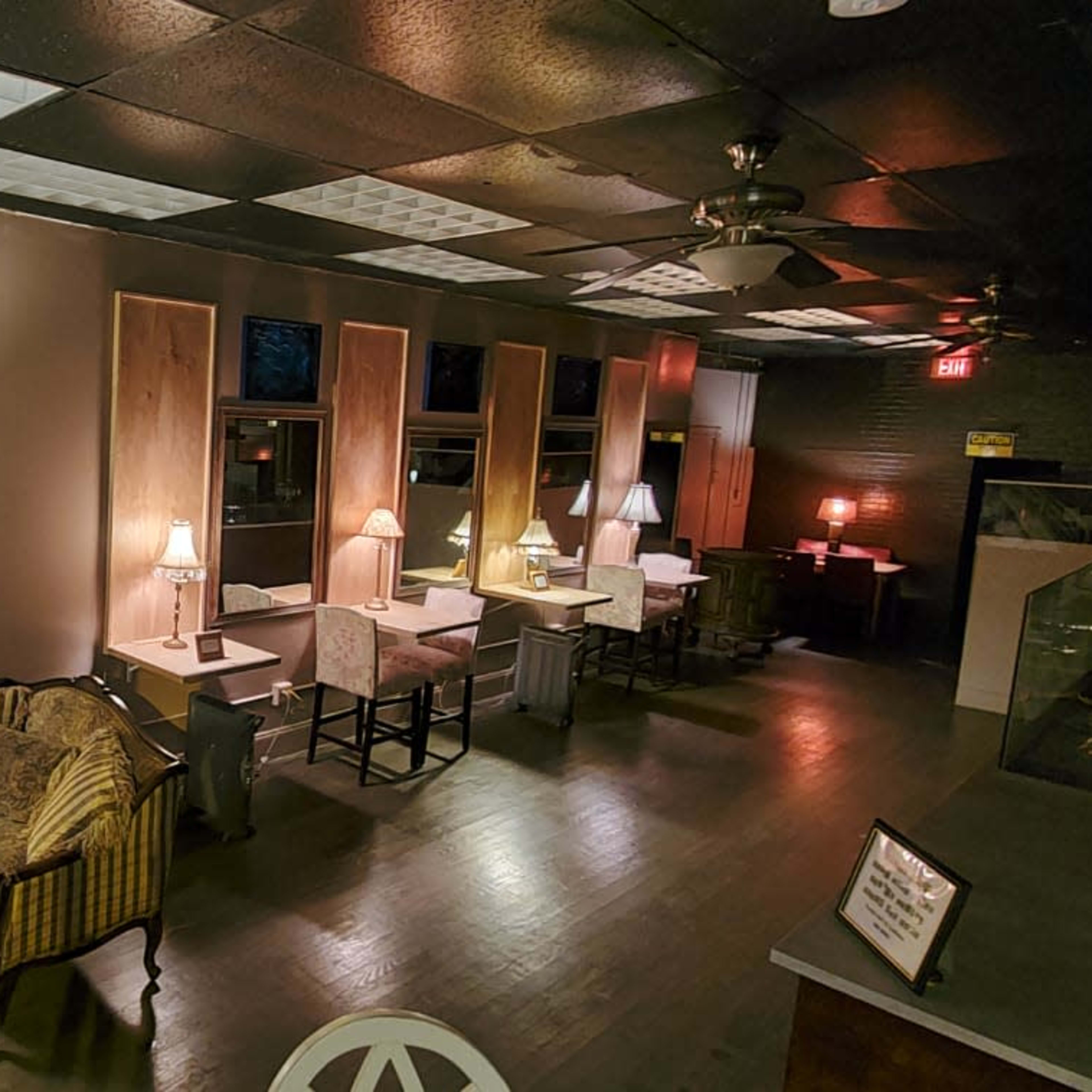A cozy cafe interior with tables, chairs, and a small stage area, illuminated by warm lamps and accented by decorative shelves and furniture.