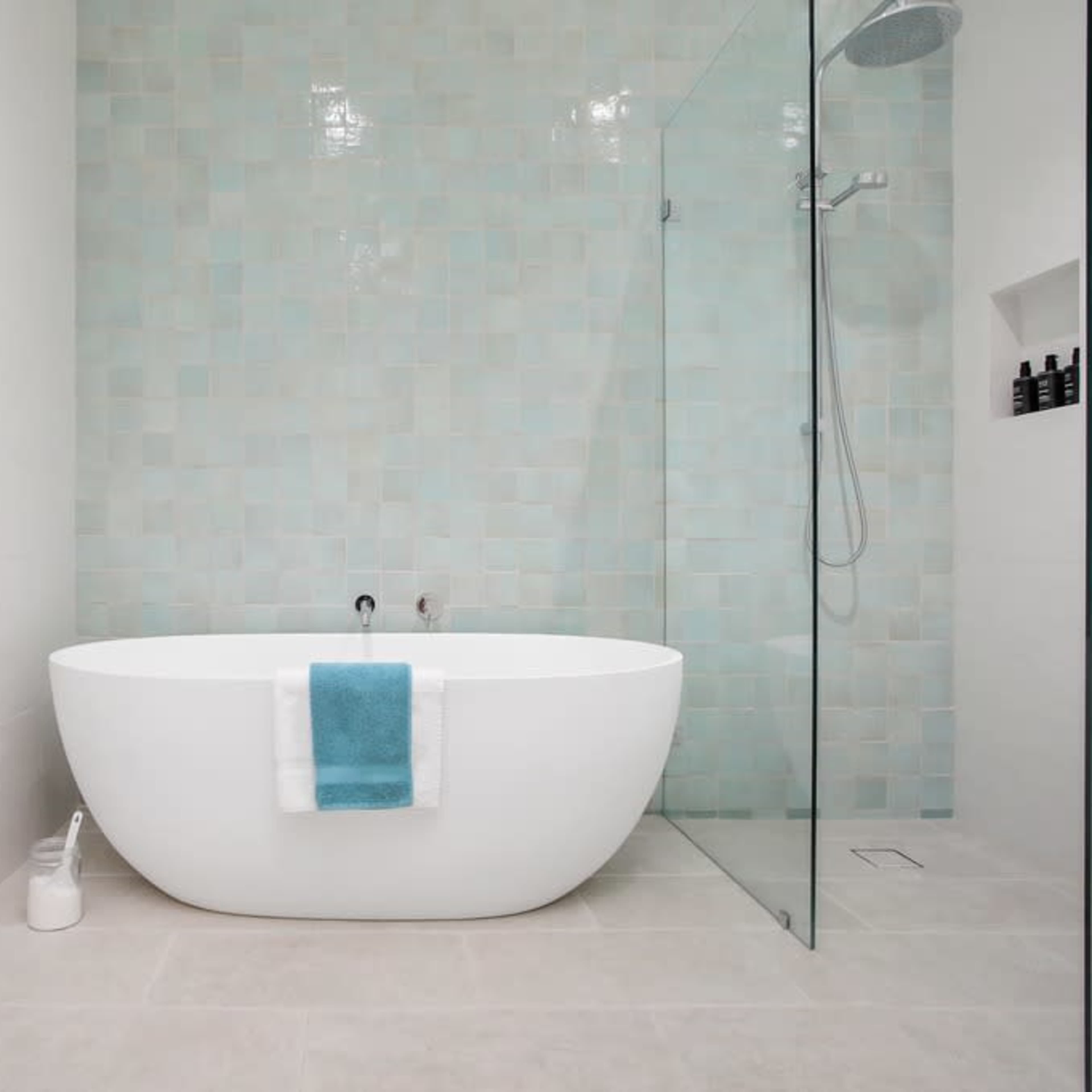 A modern bathroom featuring a freestanding white bathtub with a turquoise towel, glass shower enclosure, and light-colored tiled walls.