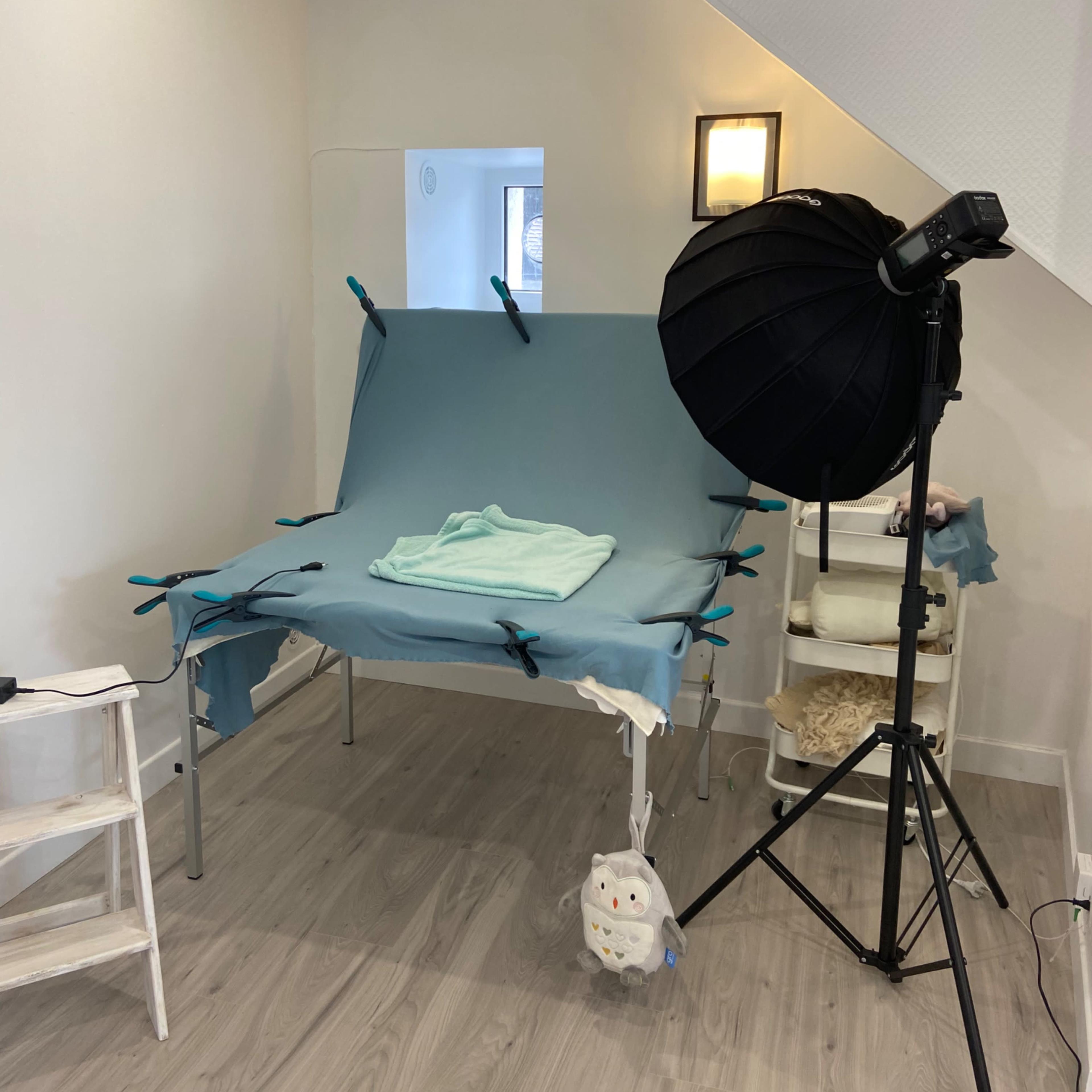 A small studio setup with a blue backdrop draped over a folding table, a photography light on a stand, and a shelf with materials in the corner.