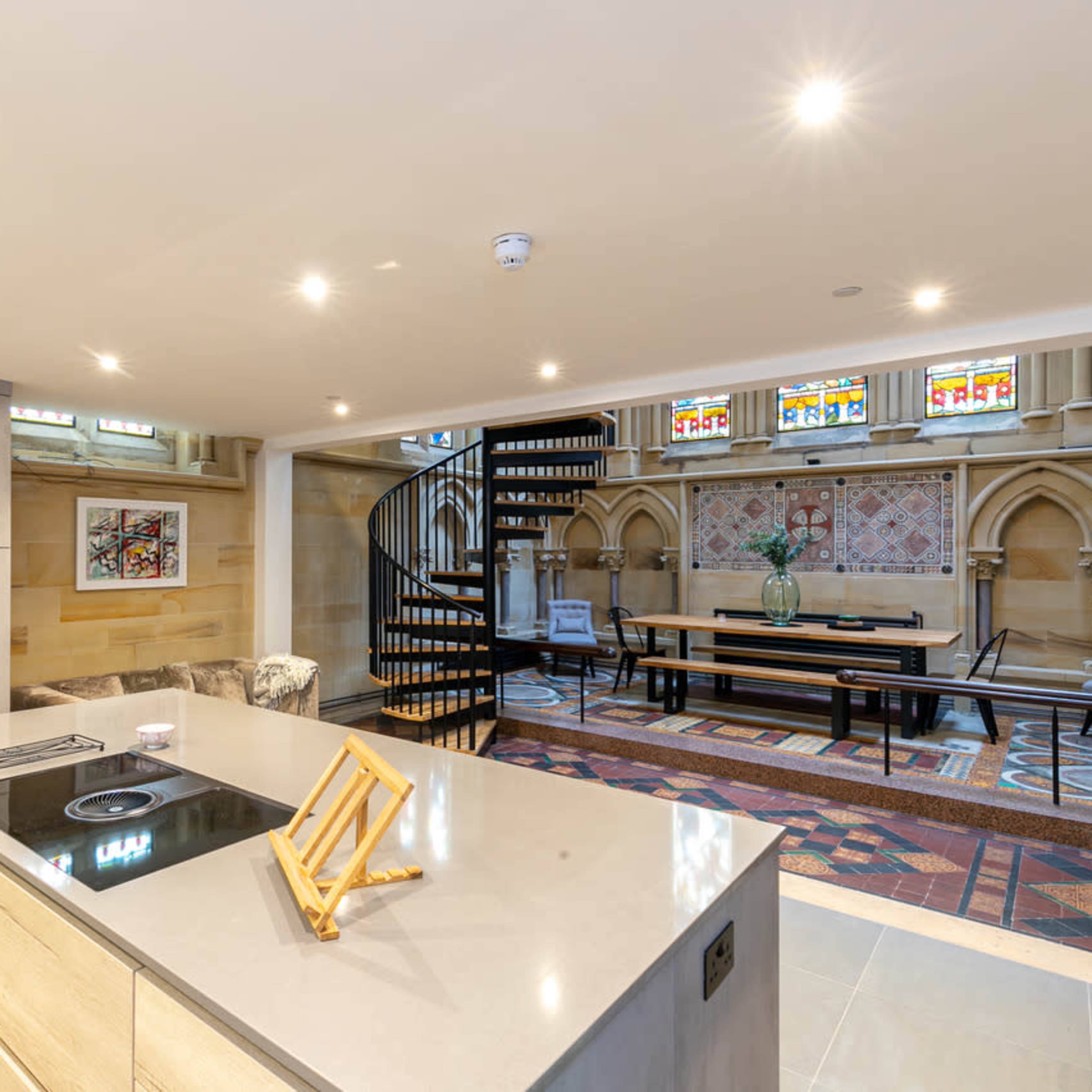 A modern kitchen with an island, featuring a spiral staircase in the background and stained glass windows illuminating the space.
