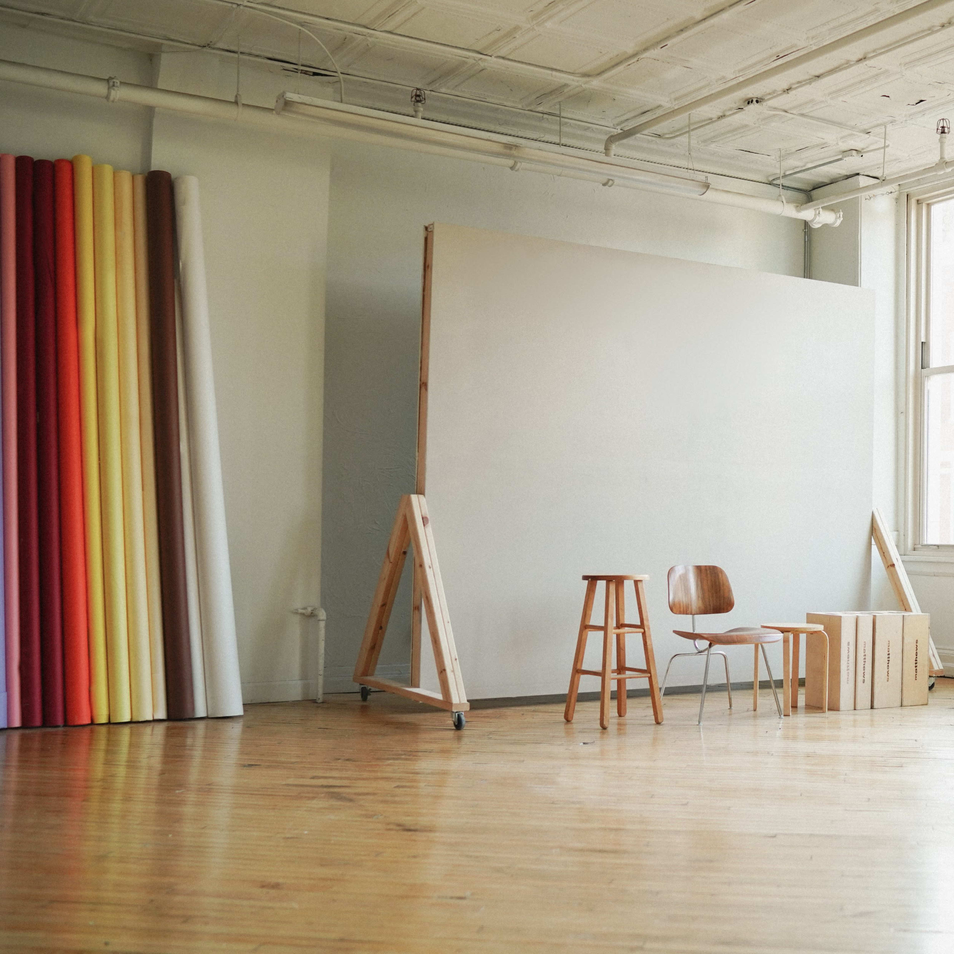 An empty studio space with a large blank wall, two wooden chairs, and rolls of colored paper against the wall.