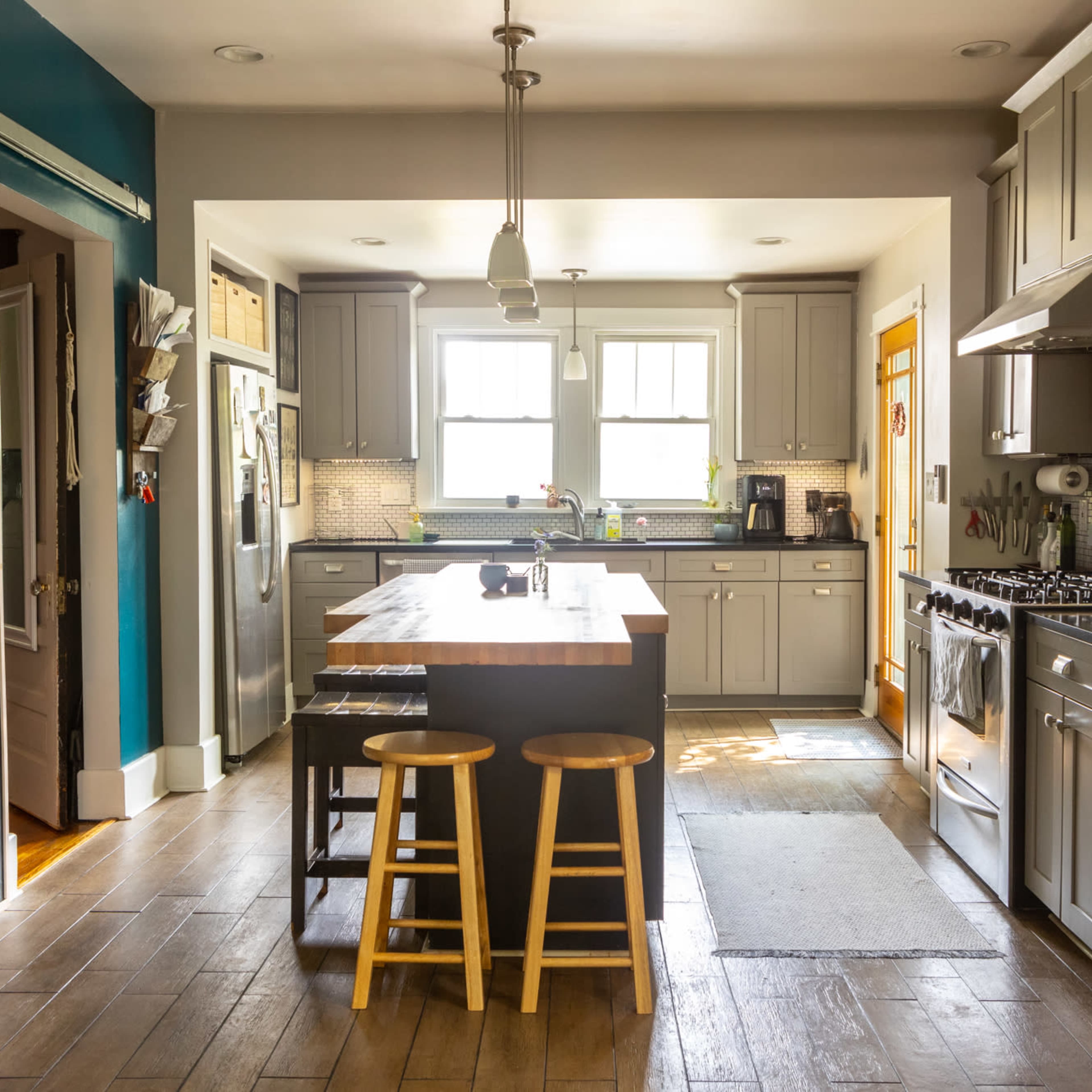 A modern kitchen with gray cabinets, an island with two stools, and stainless steel appliances, illuminated by natural light from two windows.