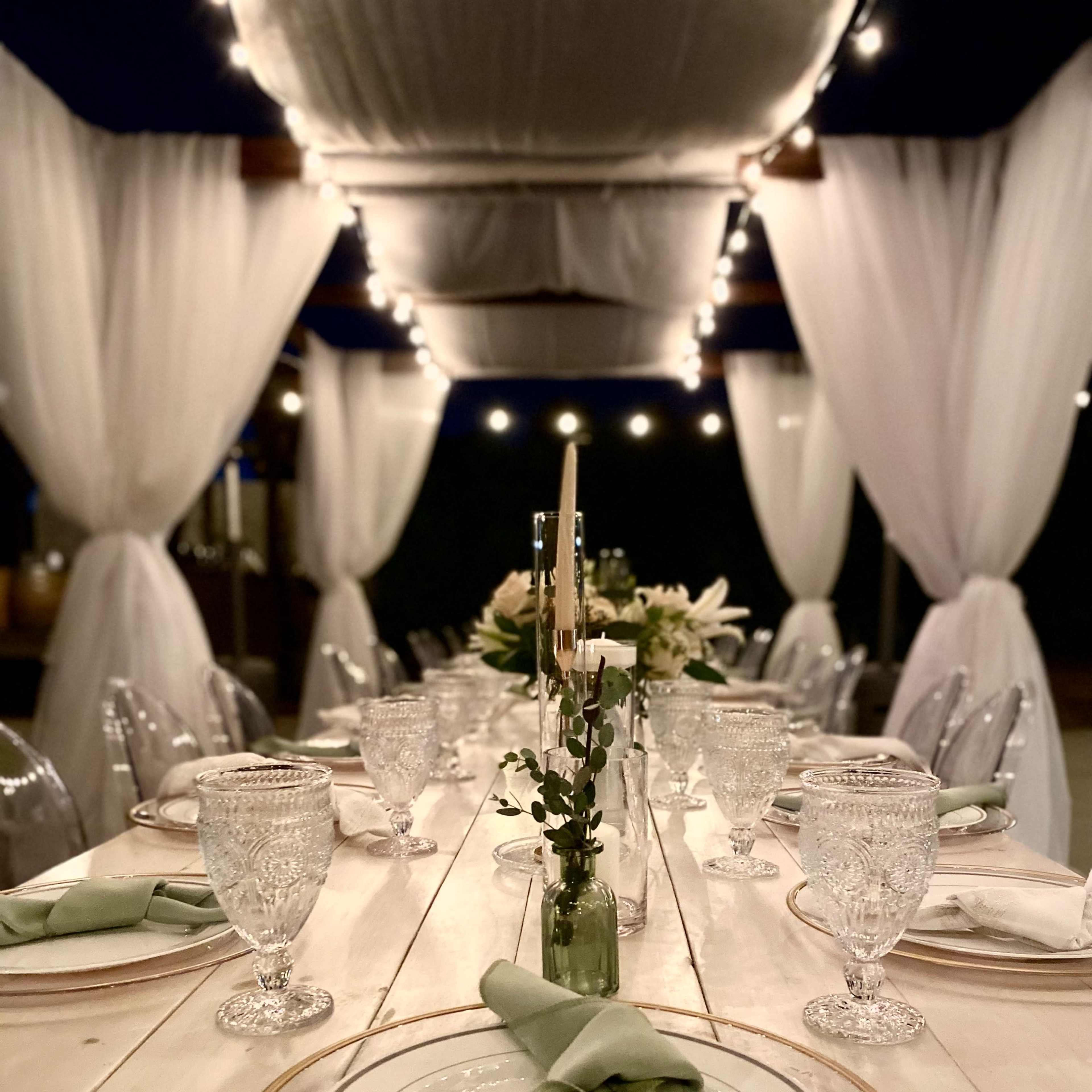 A long dining table is elegantly set with glassware, plates, and neatly folded green napkins, with draped fabric and string lights in the background.