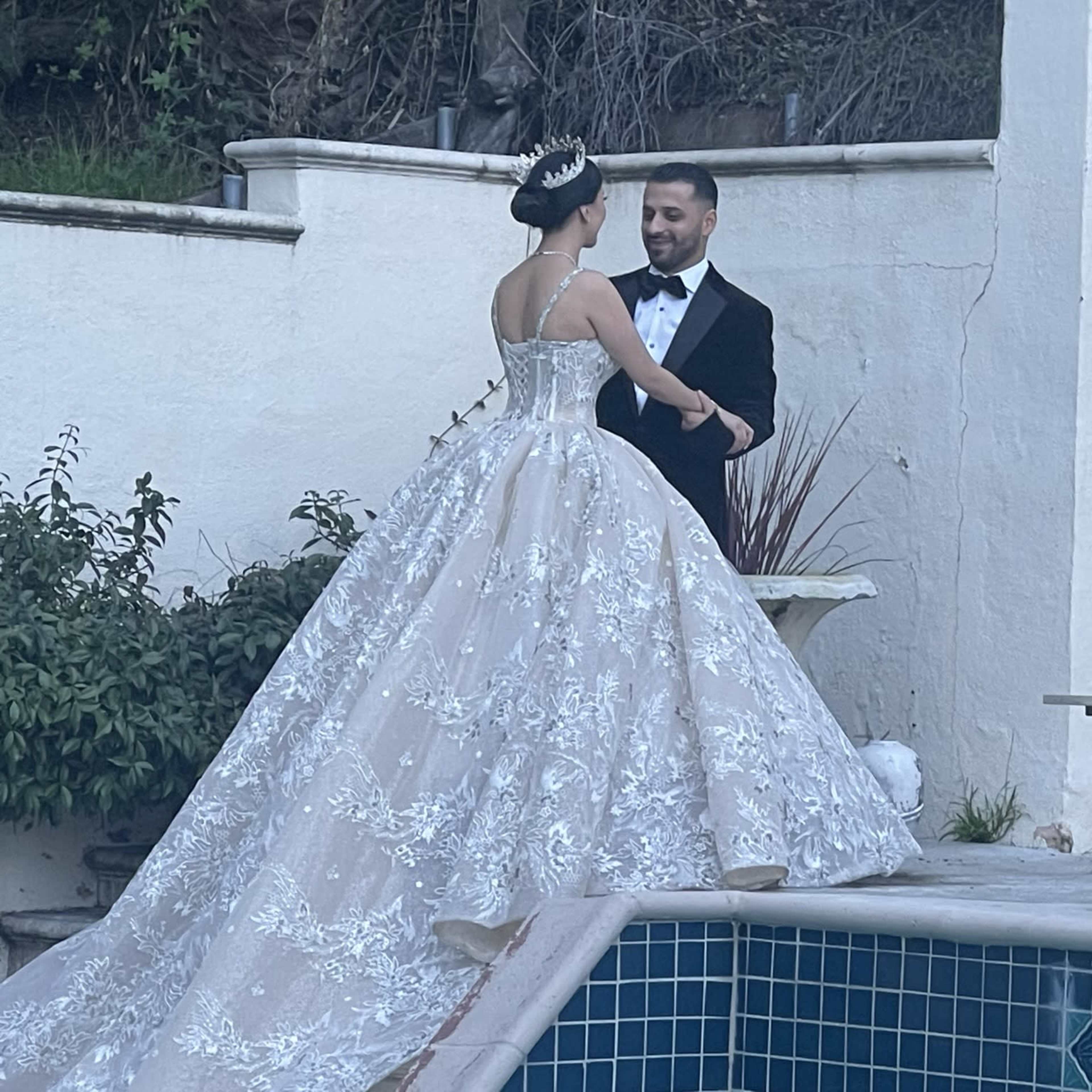A bride in a flowing gown and a groom in a tuxedo share a moment near a poolside setting.