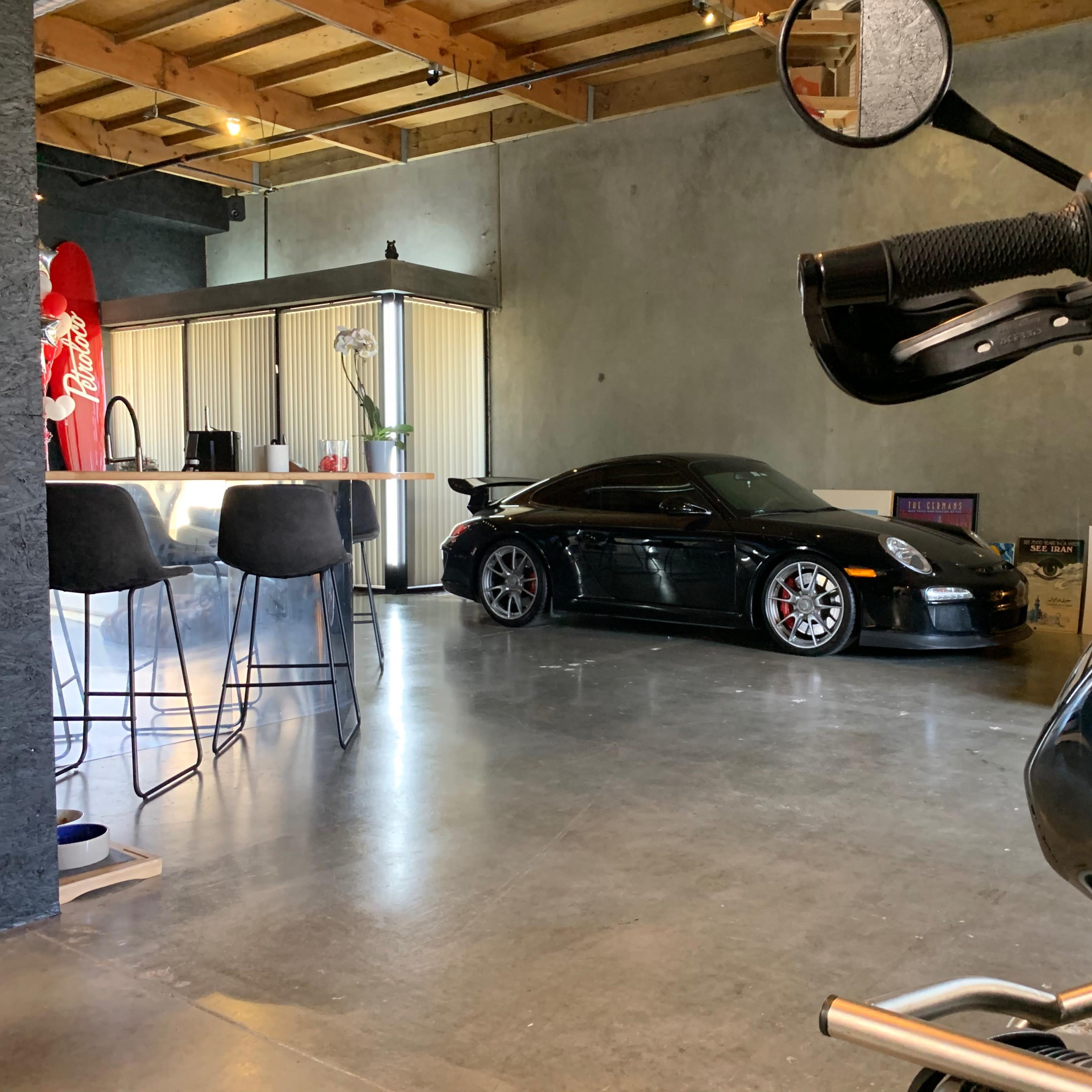 A black sports car is parked in a modern garage featuring concrete walls and minimalistic decor, with a motorcycle visible in the foreground.