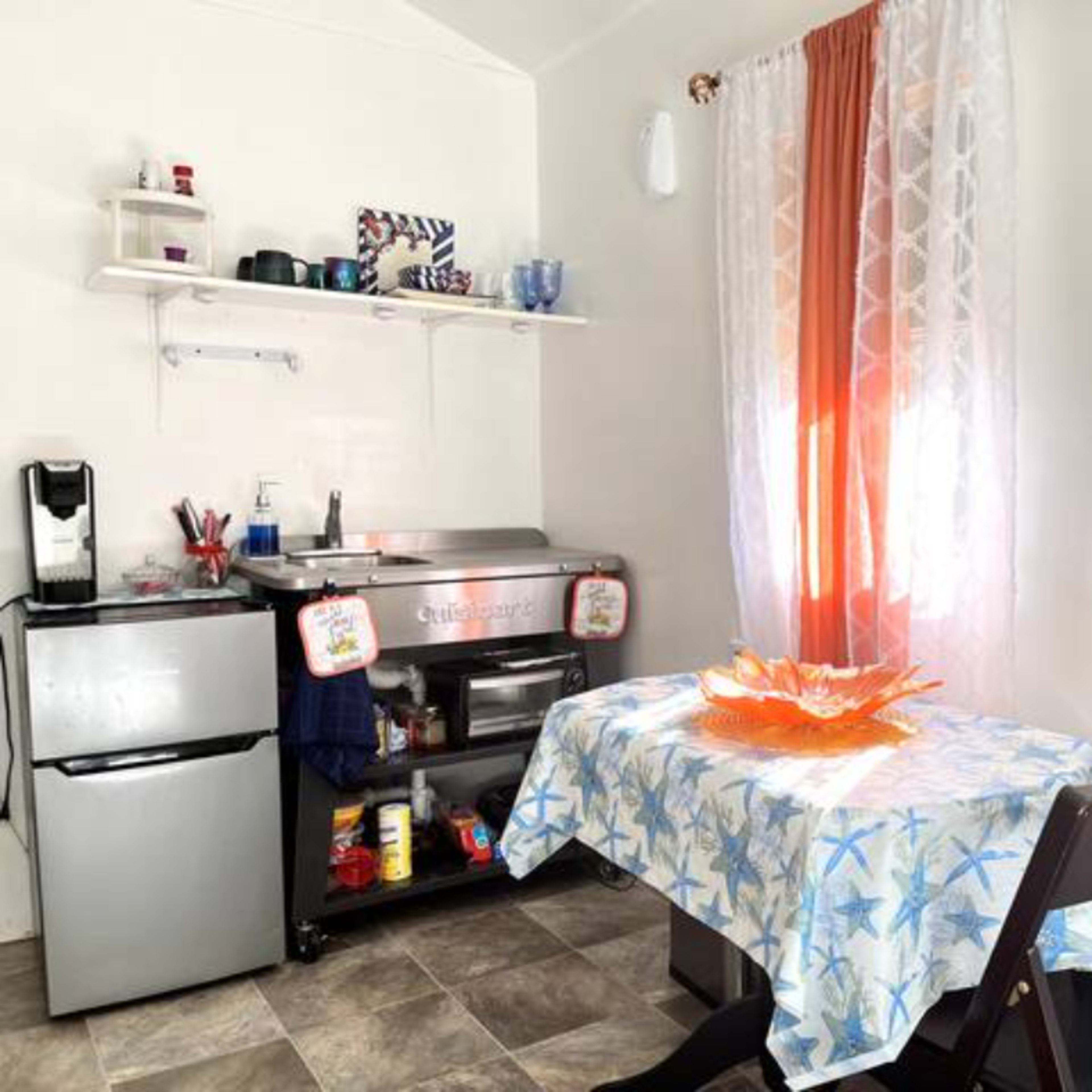 A small kitchen area features a table covered with a blue and white tablecloth, a fruit bowl in the center, and kitchen appliances and shelves against the wall.