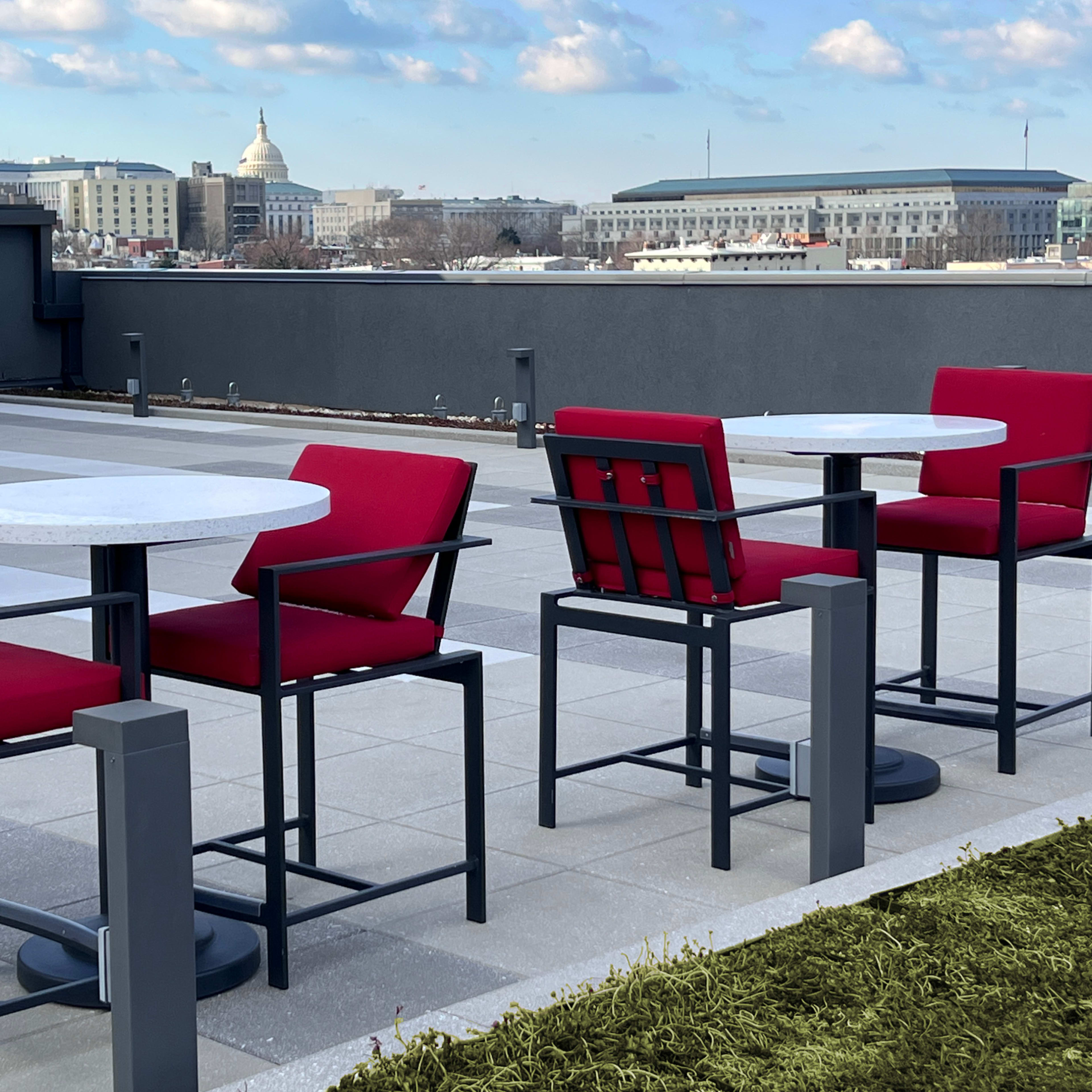 Two red cushioned chairs are positioned at white tables on a rooftop terrace, with a view of buildings and the Capitol dome in the background.
