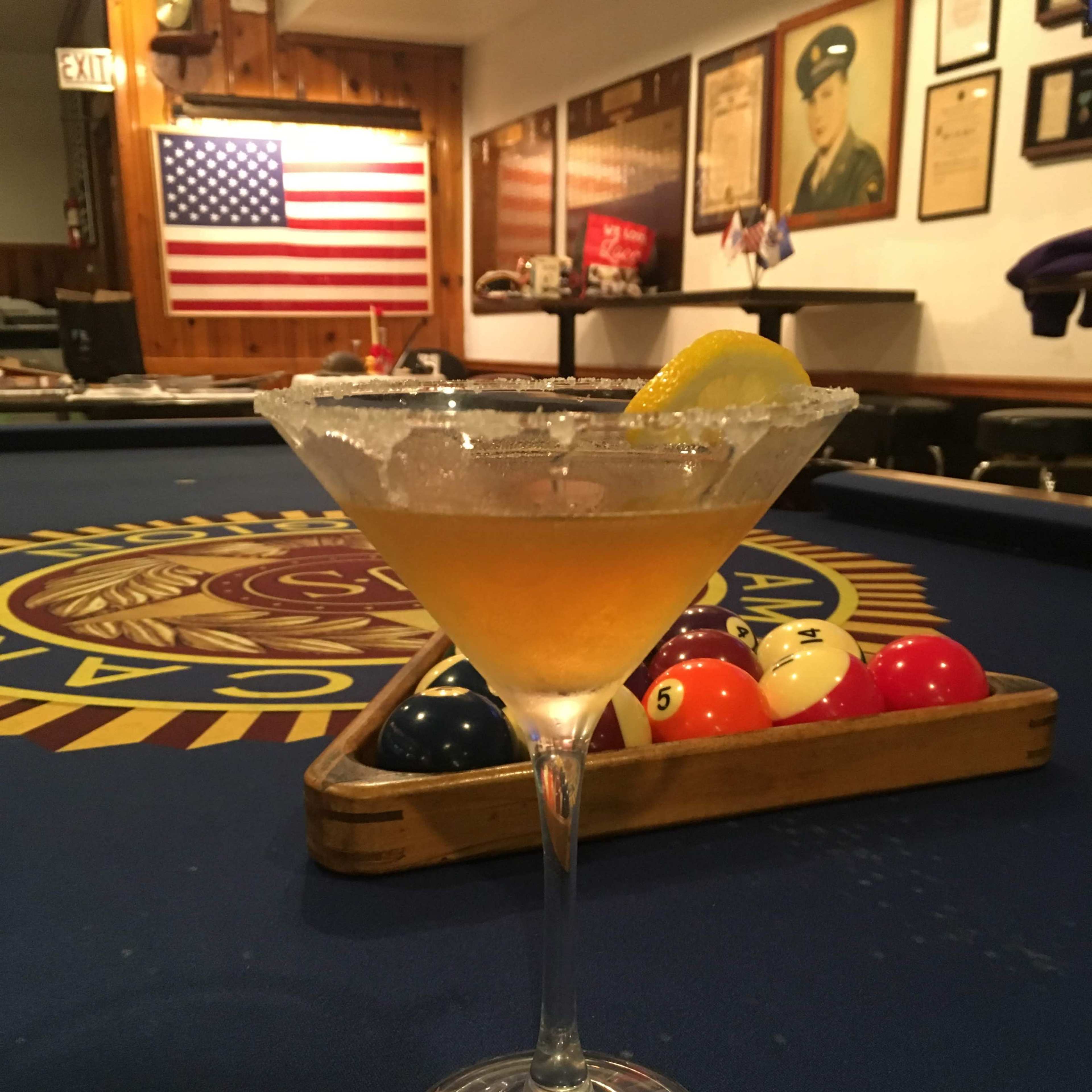 A cocktail with a lemon wedge and a sugared rim is placed in front of a pool table in a bar decorated with an American flag and military memorabilia.