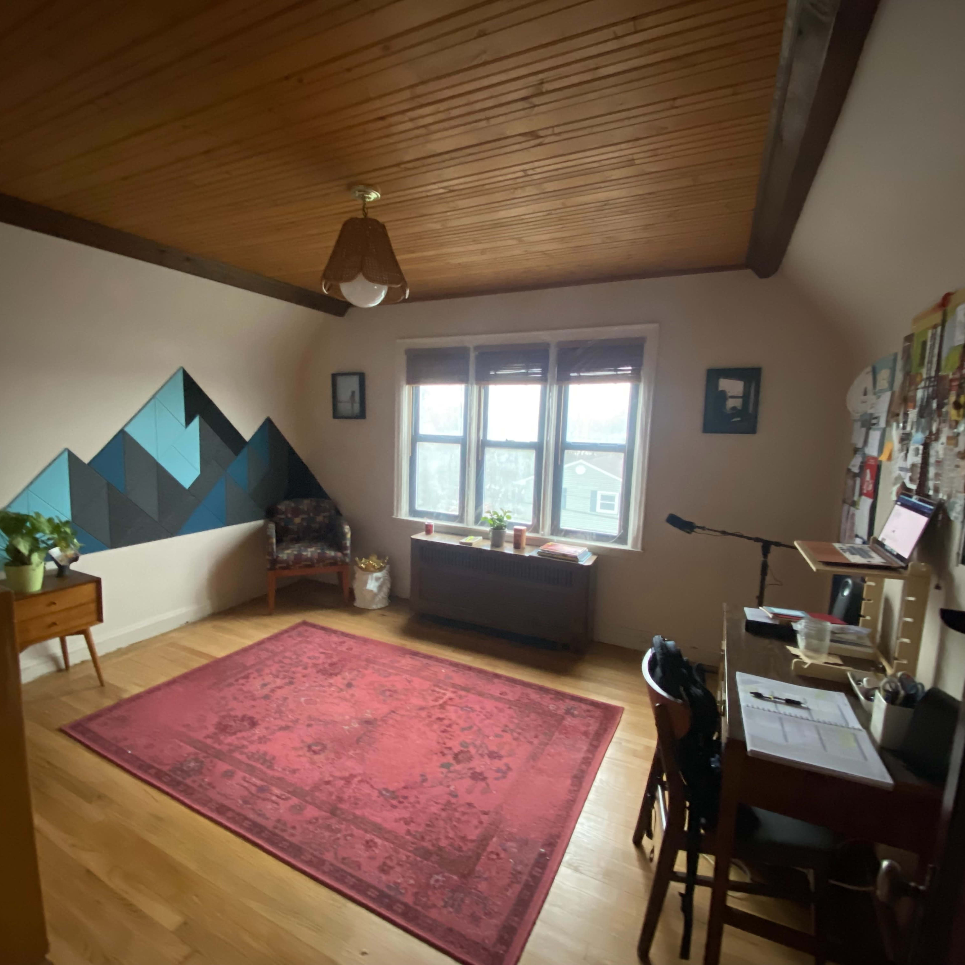 A spacious room with wooden beams on the ceiling, a patterned rug on the floor, a desk with a laptop, and a small seating area by the windows.